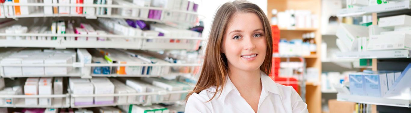 Un farmacéutico sonriente se encuentra en una farmacia con estantes repletos de cajas de medicamentos al fondo.