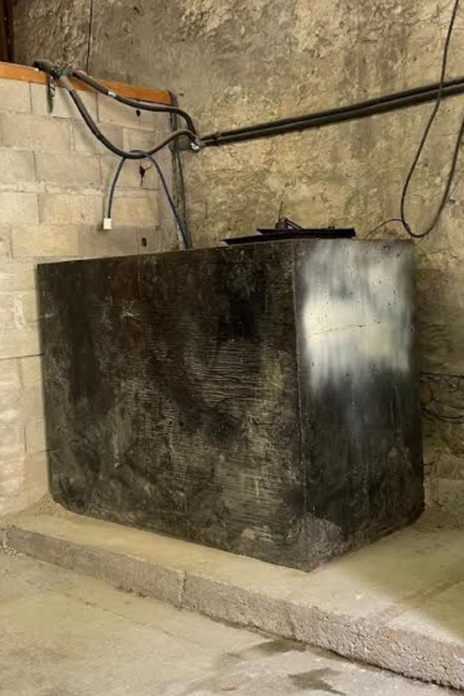 Réservoir rectangulaire noir posé sur un bloc de béton, adossé à un mur de briques et de pierres patiné, avec des câbles aériens.