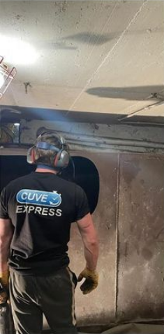 Un homme portant des protections auditives et des gants de travail perce une structure métallique. L'inscription CUVE EXPRESS figure au dos de son t-shirt.