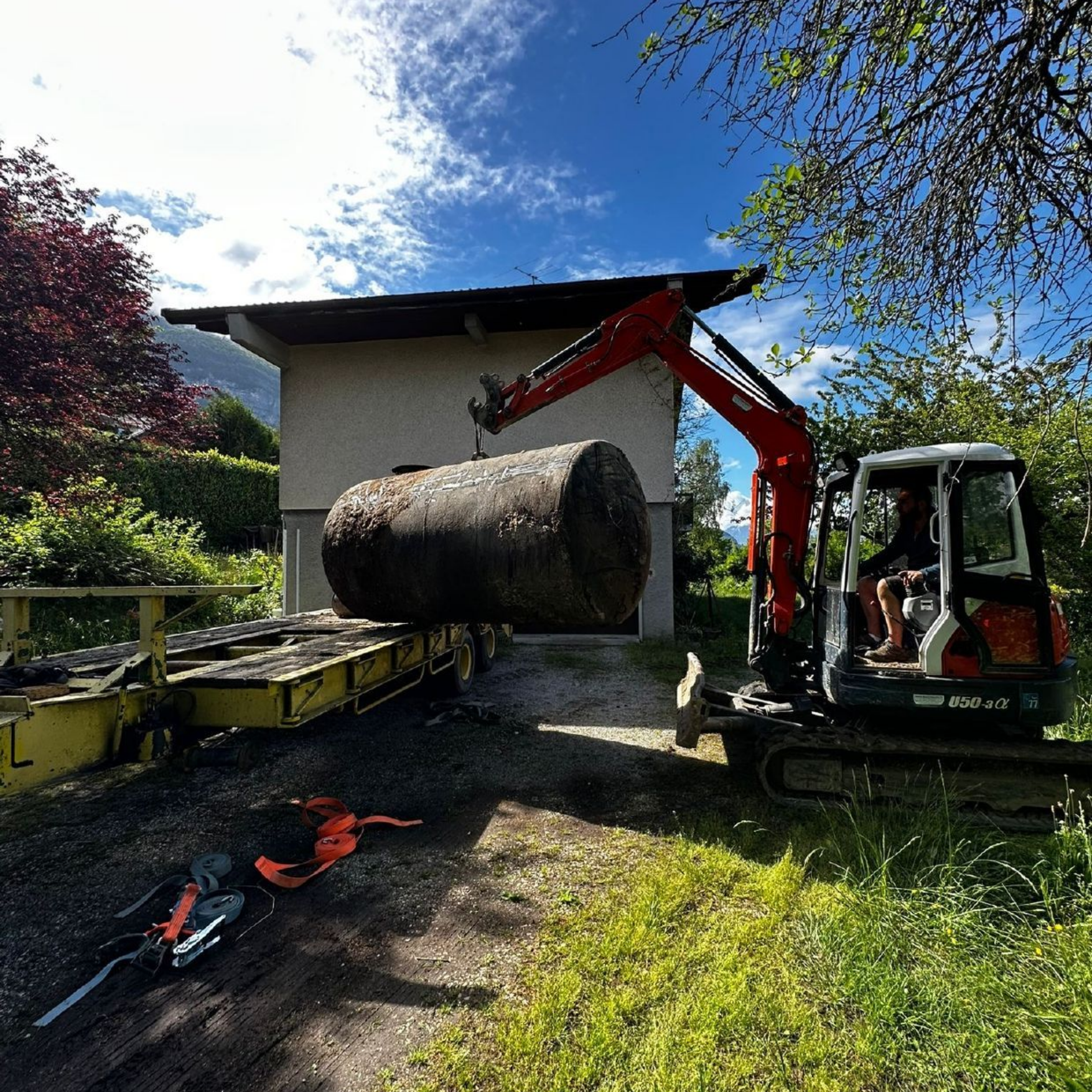 Une minipelle charge un gros cylindre de béton sur une remorque devant un bâtiment par une journée ensoleillée.