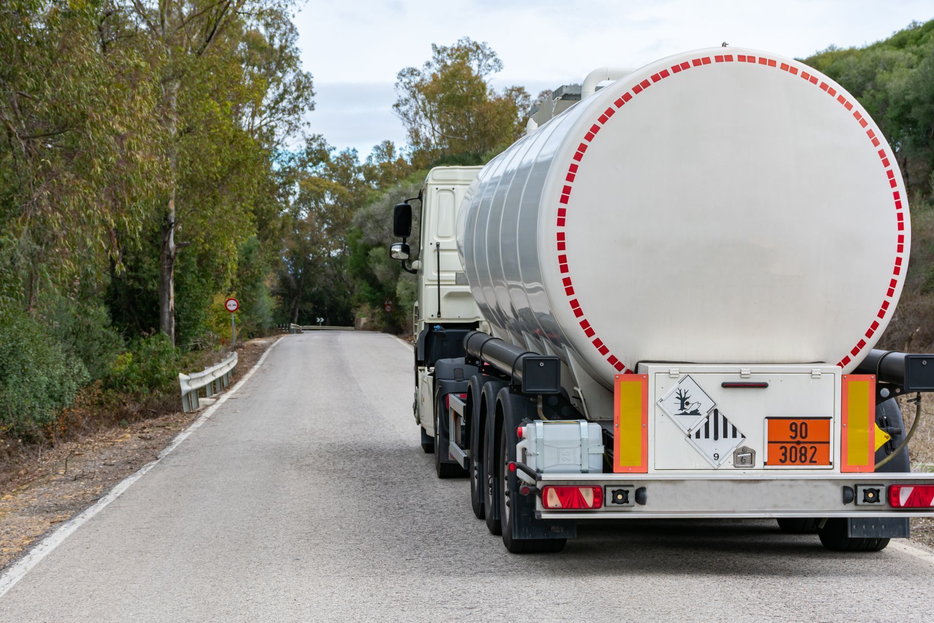 Camion-citerne sur une route étroite ; citerne blanche, panneau de danger jaune/orange, arbres.