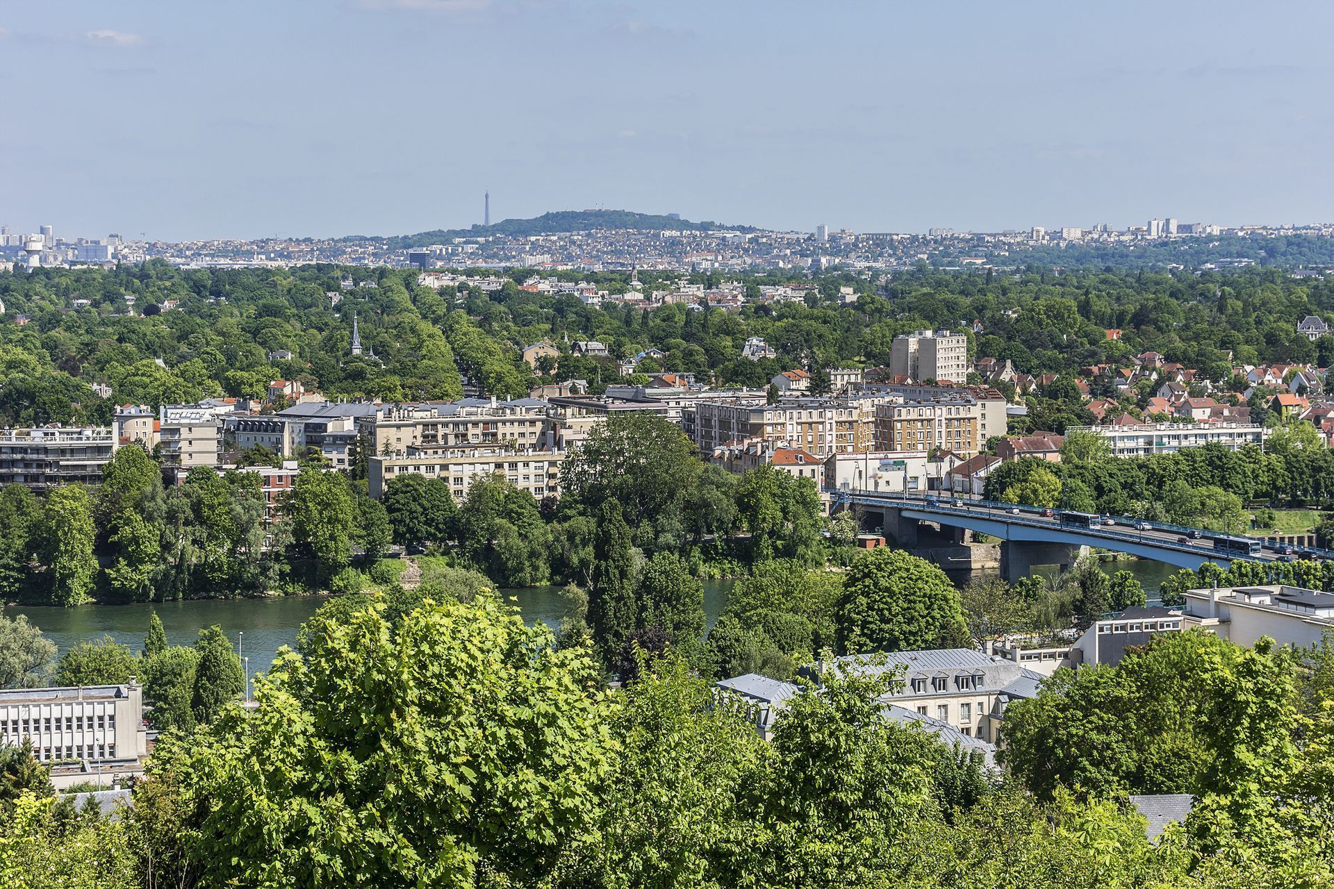 Paysage urbain avec bâtiments, arbres et eau sous un ciel bleu, colline au loin avec une tour.