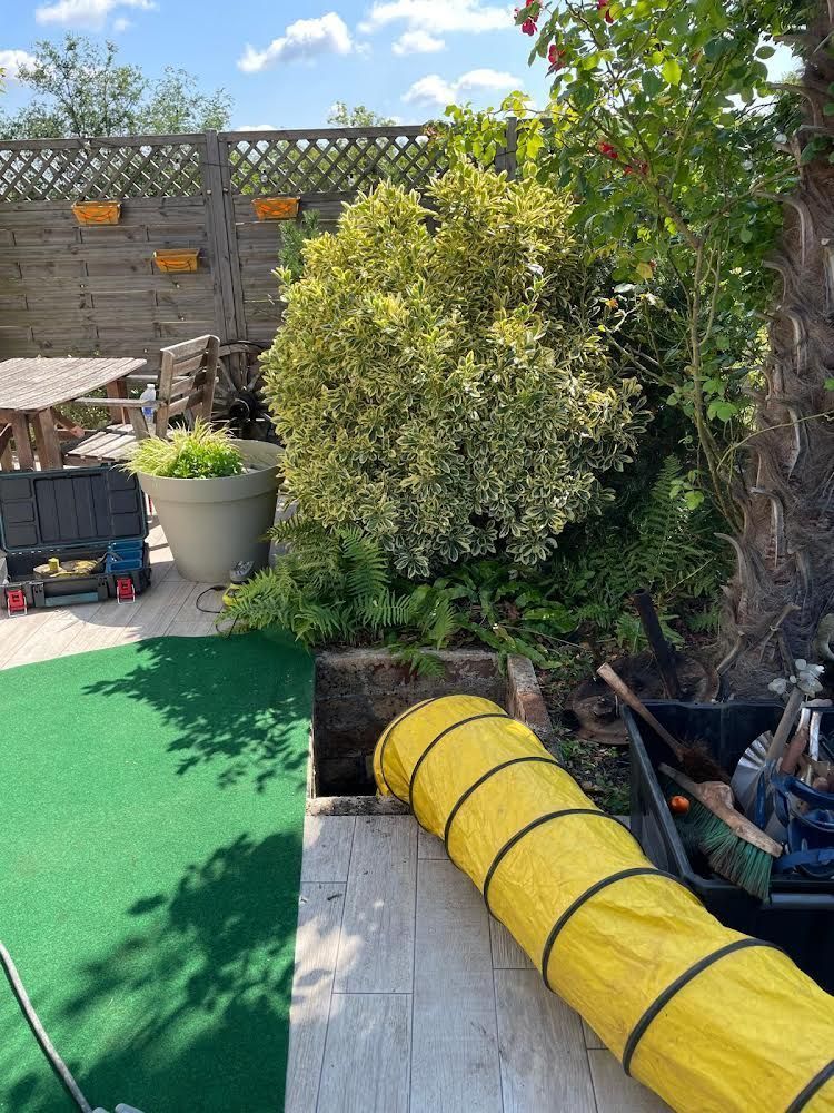 Un tuyau jaune sort d'un trou carré sombre dans une terrasse pavée, avec un jardin en arrière-plan.