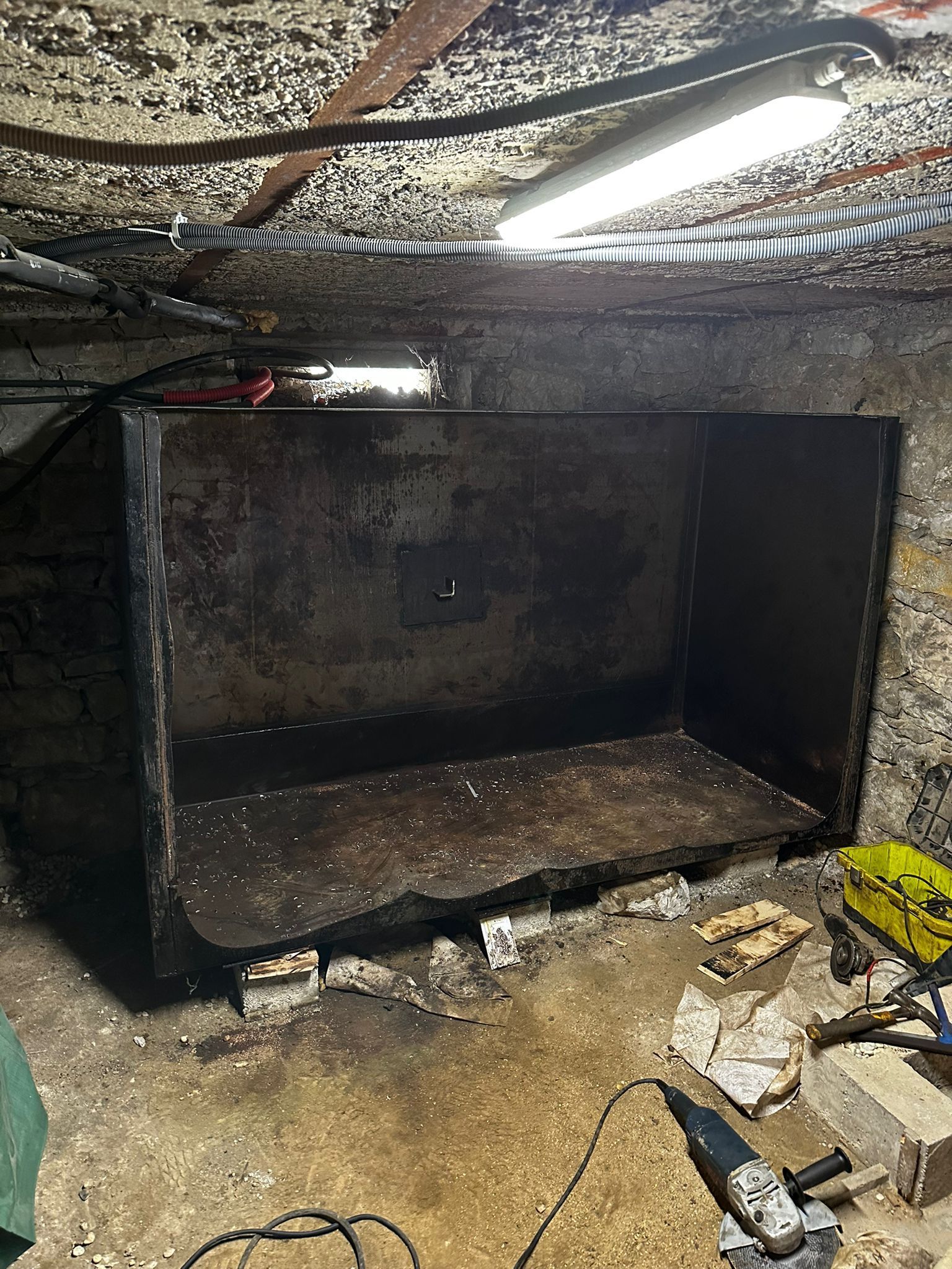 Un grand conteneur métallique sombre se trouve dans un sous-sol encombré. Les murs et le plafond sont vieux et délabrés.