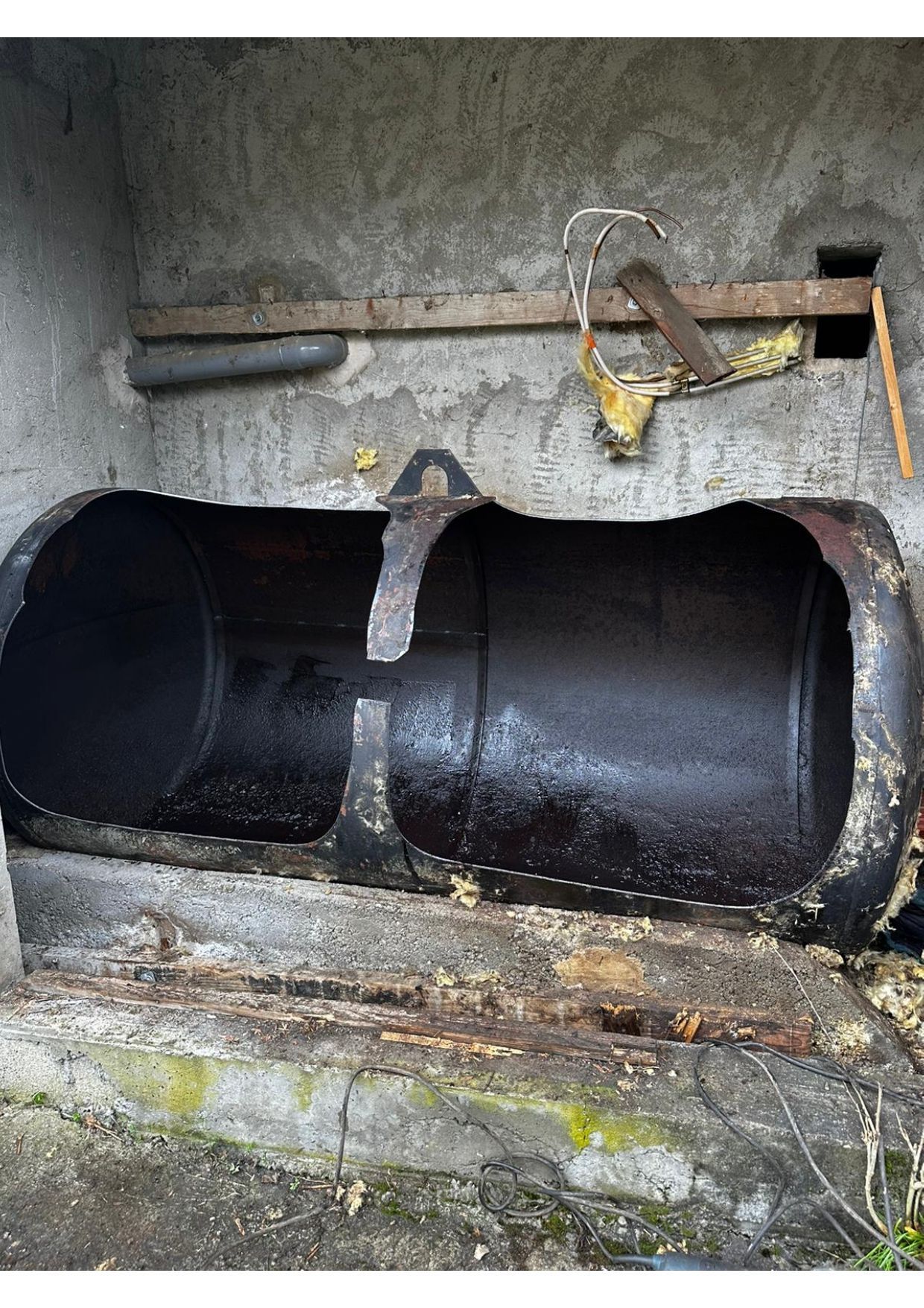 Un tuyau métallique sombre, ouvert, est scellé dans du béton. Une poutre en bois et des outils métalliques rouillés se trouvent au-dessus.