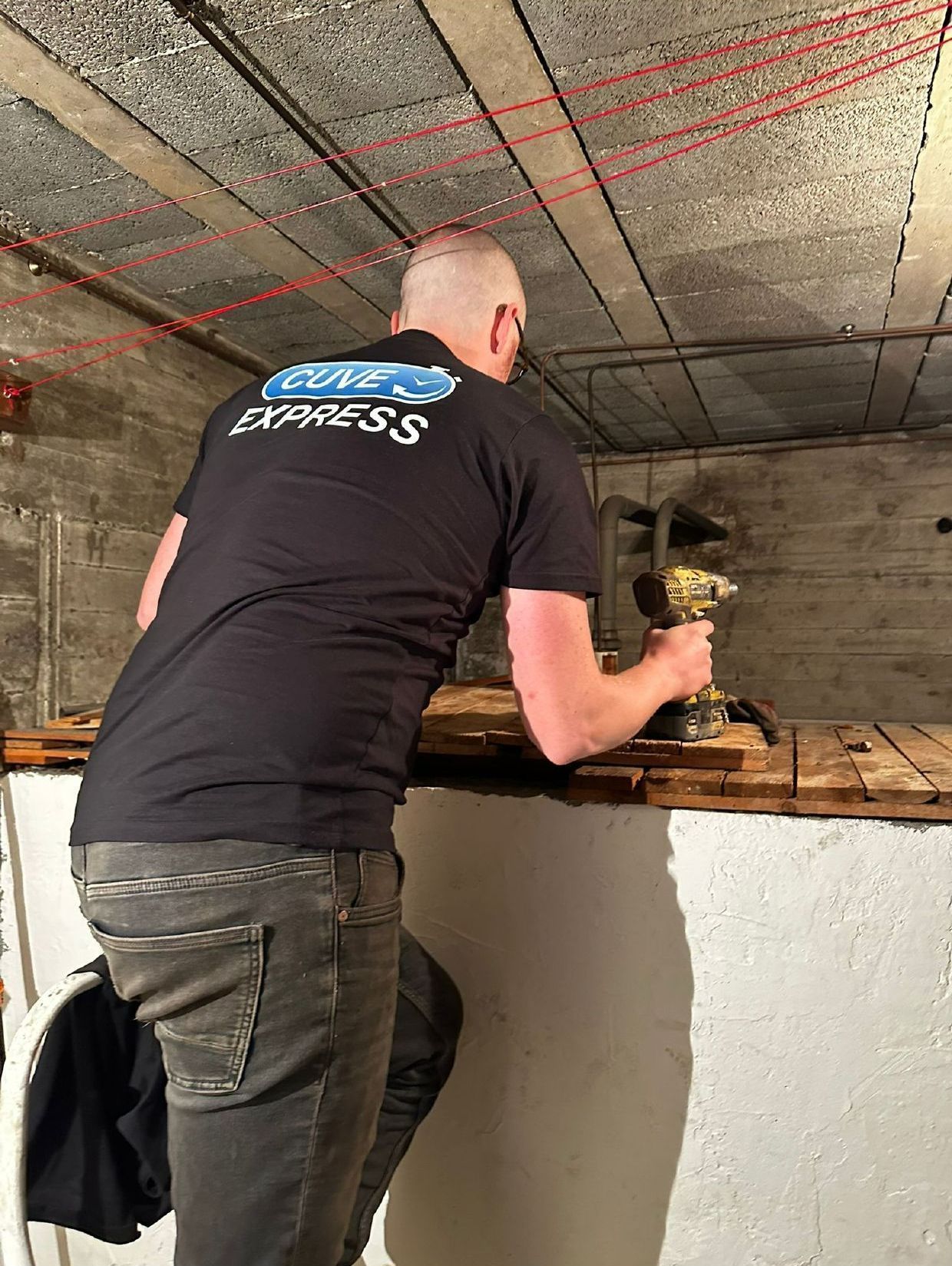 Personne en chemise noire, travaillant avec un outil sur une surface en bois dans un sous-sol, lignes rouges au-dessus de la tête.