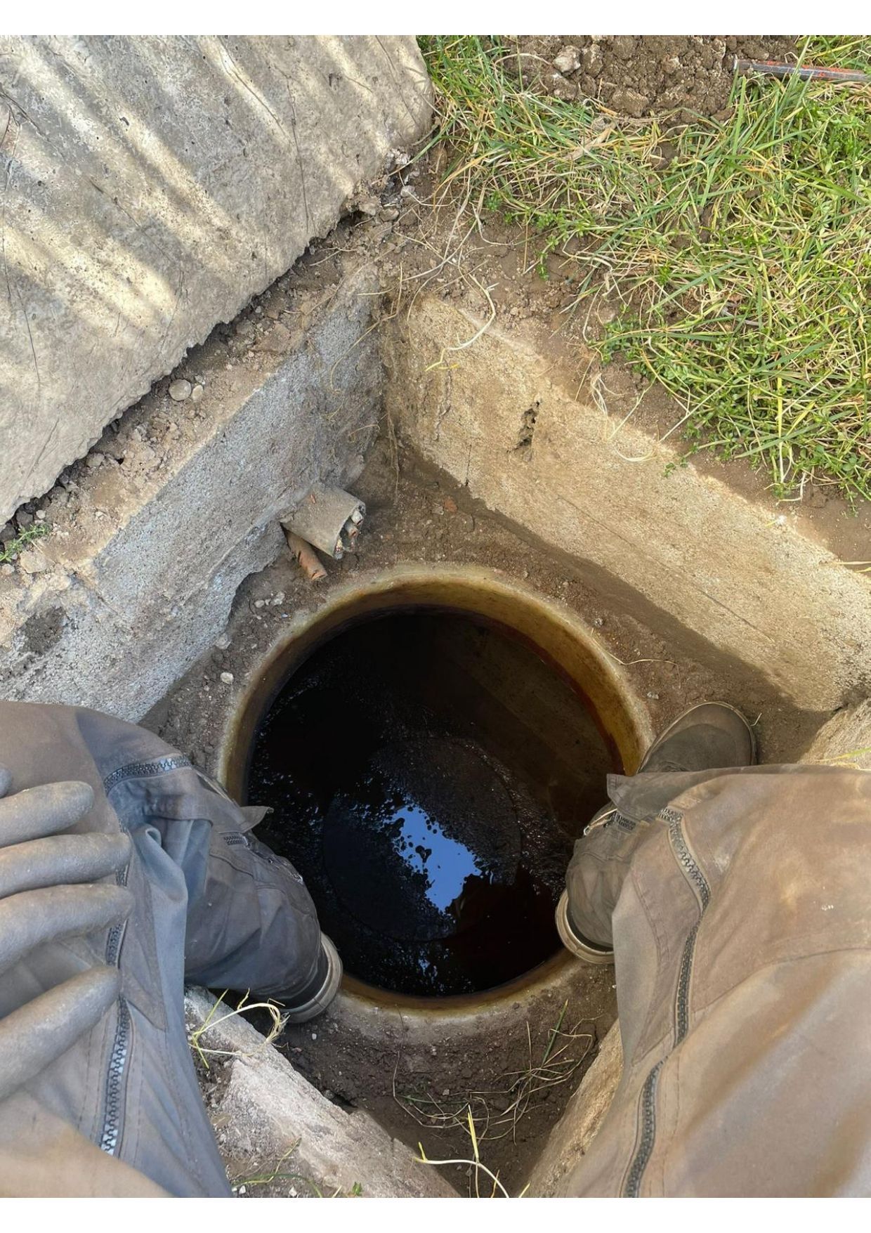 Une personne se tient au-dessus d'une bouche d'égout circulaire, ouverte et sombre, entourée de béton et d'herbe.