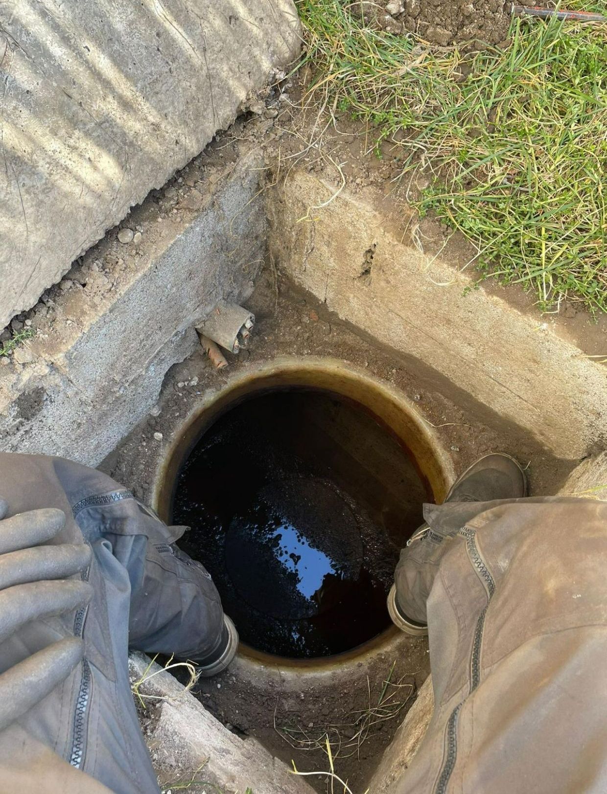 Une personne regarde à l'intérieur d'une bouche d'égout sombre et circulaire, entourée de béton et d'herbe.