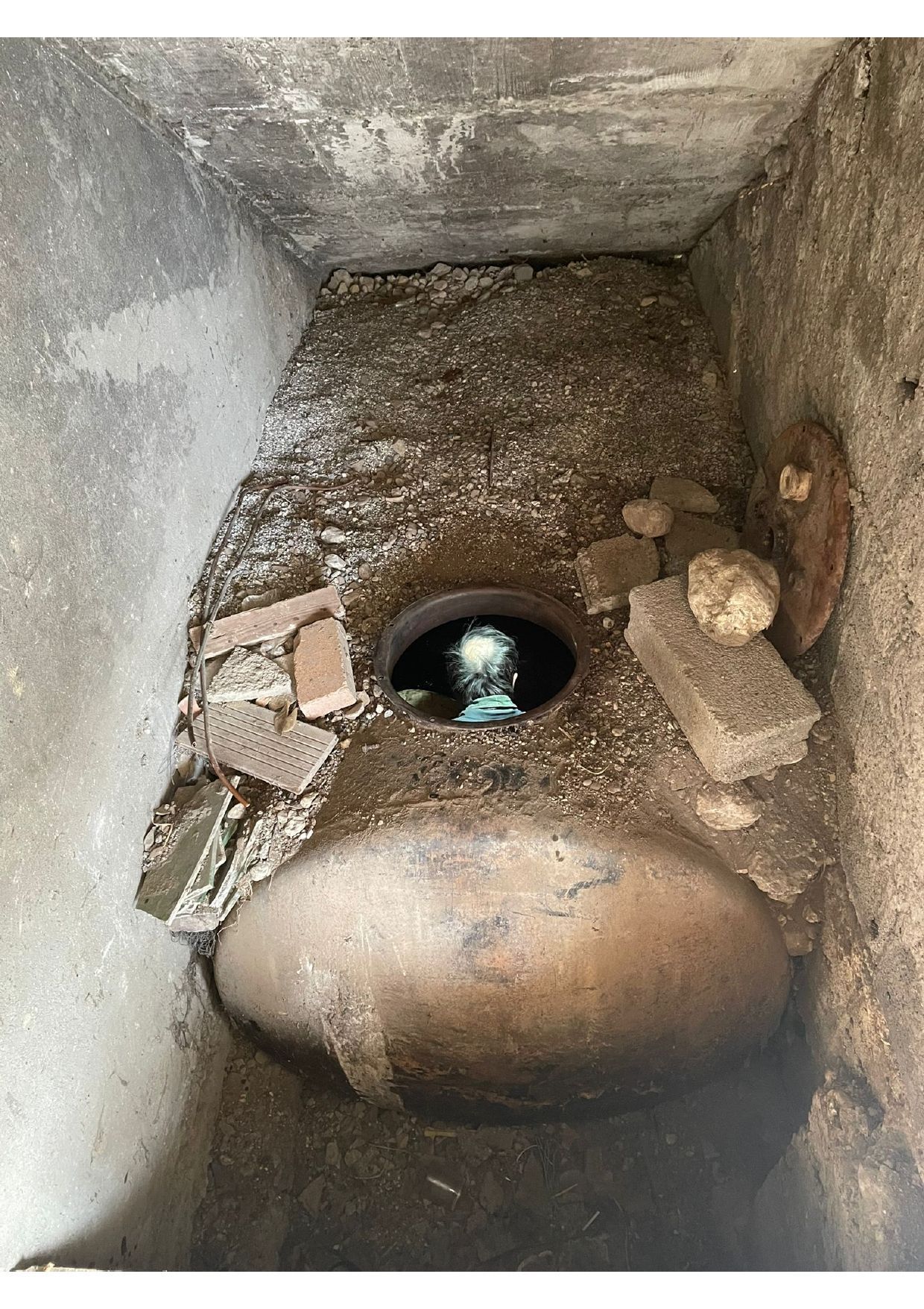 À l'intérieur d'une fosse en béton : un drain circulaire à ciel ouvert, entouré de briques cassées et de terre.