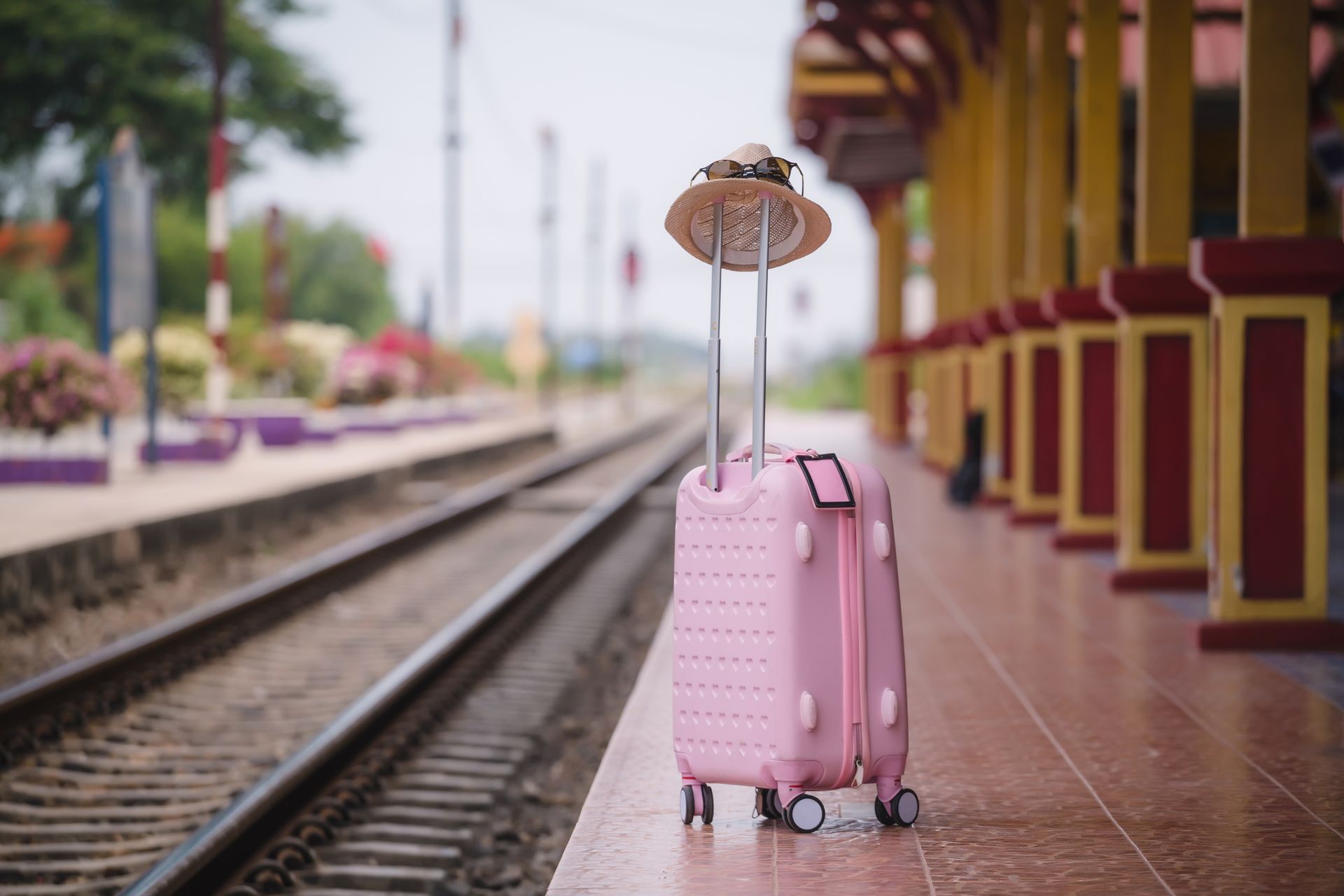 Une valise rose, sur laquelle est posé un chapeau de soleil appuyé contre sa poignée déployée, se trouve sur le quai d'une gare.