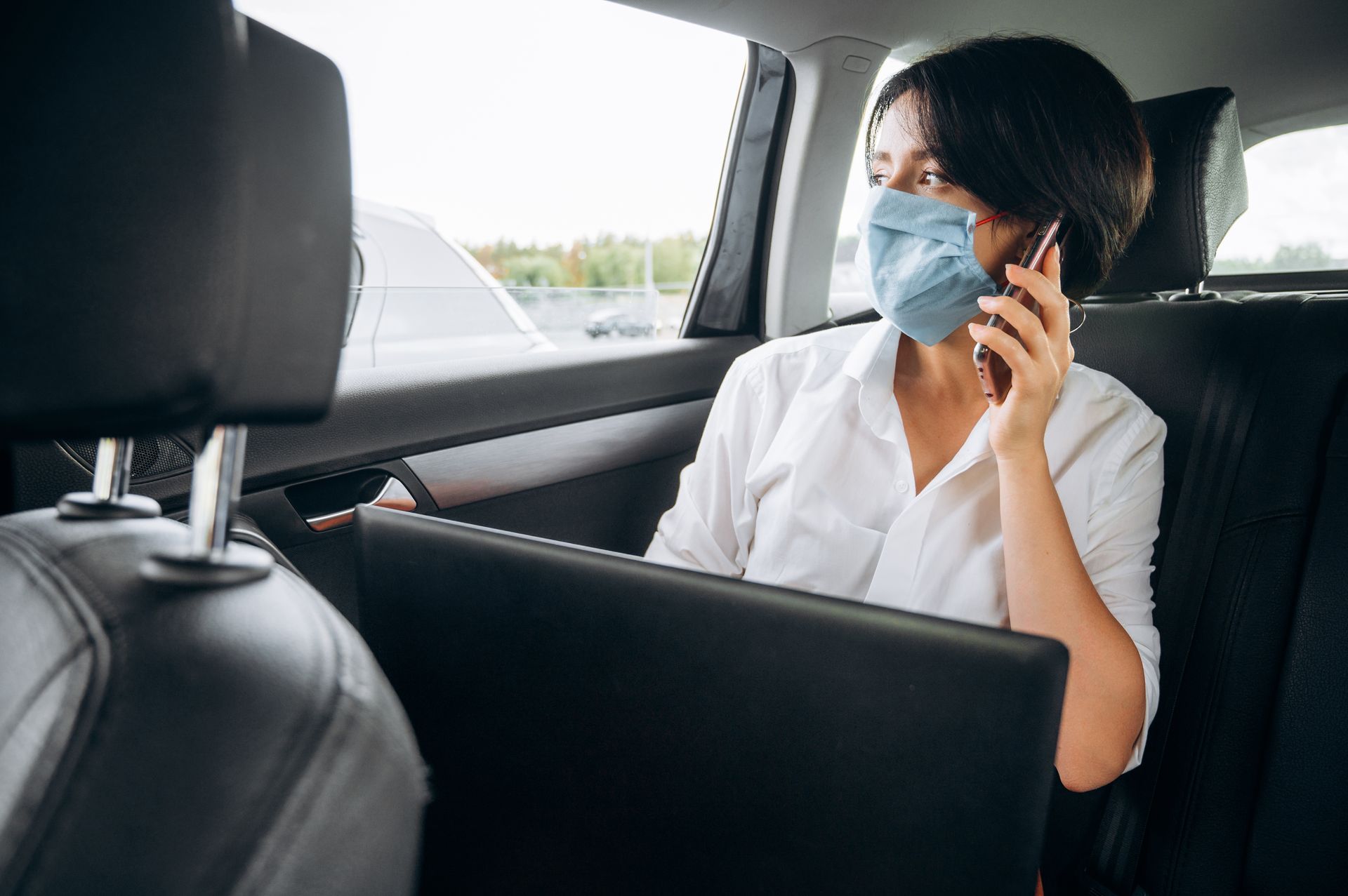 Une personne portant un masque est assise à l'arrière d'une voiture, travaillant sur un ordinateur portable tout en parlant au téléphone portable.