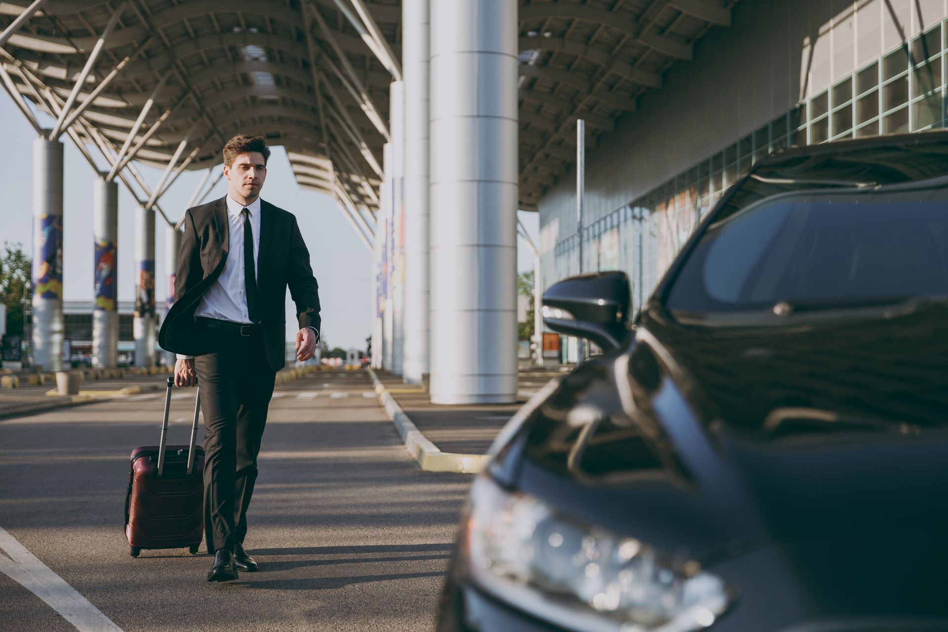 Un homme d'affaires en costume passe avec une valise devant une voiture garée à l'extérieur d'un terminal d'aéroport moderne.