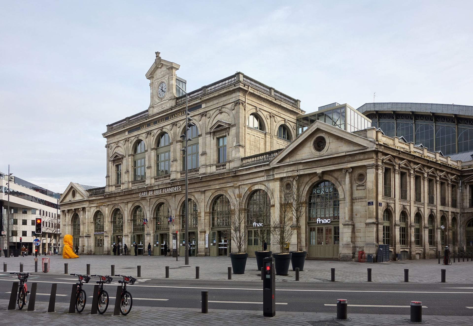 Un bâtiment historique beige à plusieurs étages, avec une colonnade voûtée et une passerelle vitrée sous un ciel bleu limpide.