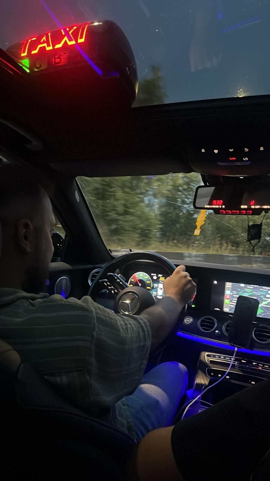 Un chauffeur conduit un taxi de nuit, avec un panneau lumineux rouge indiquant qu'il s'agit d'un taxi, visible à travers le pare-brise.