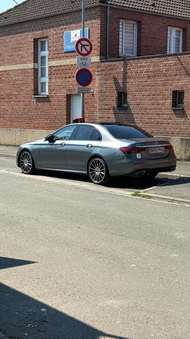 Une berline Mercedes-Benz argentée garée sur une rue asphaltée devant un bâtiment en briques rouges avec des panneaux de signalisation.