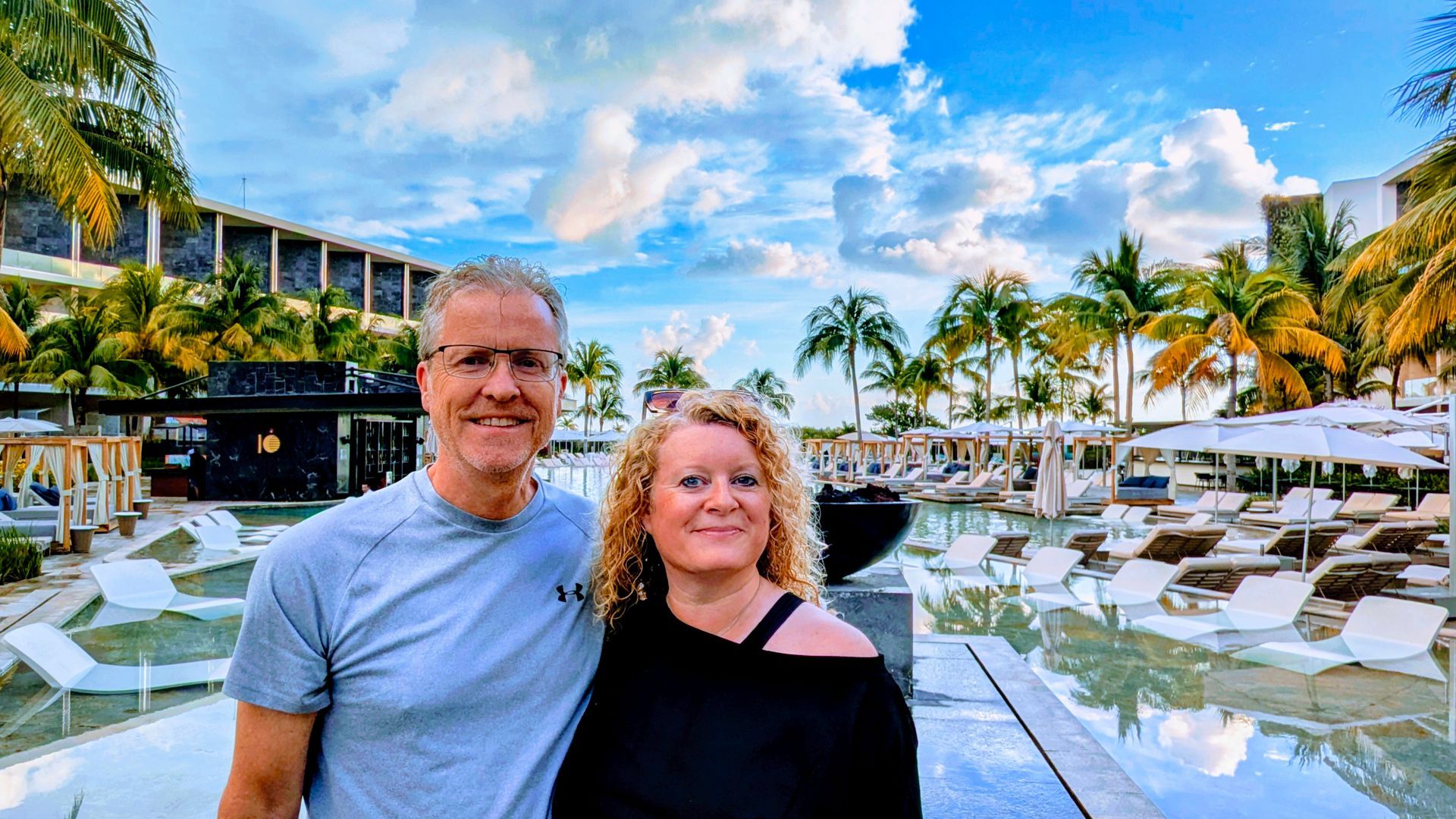 Couple smiling near a pool at a resort, with palm trees and a cloudy sky in the background.