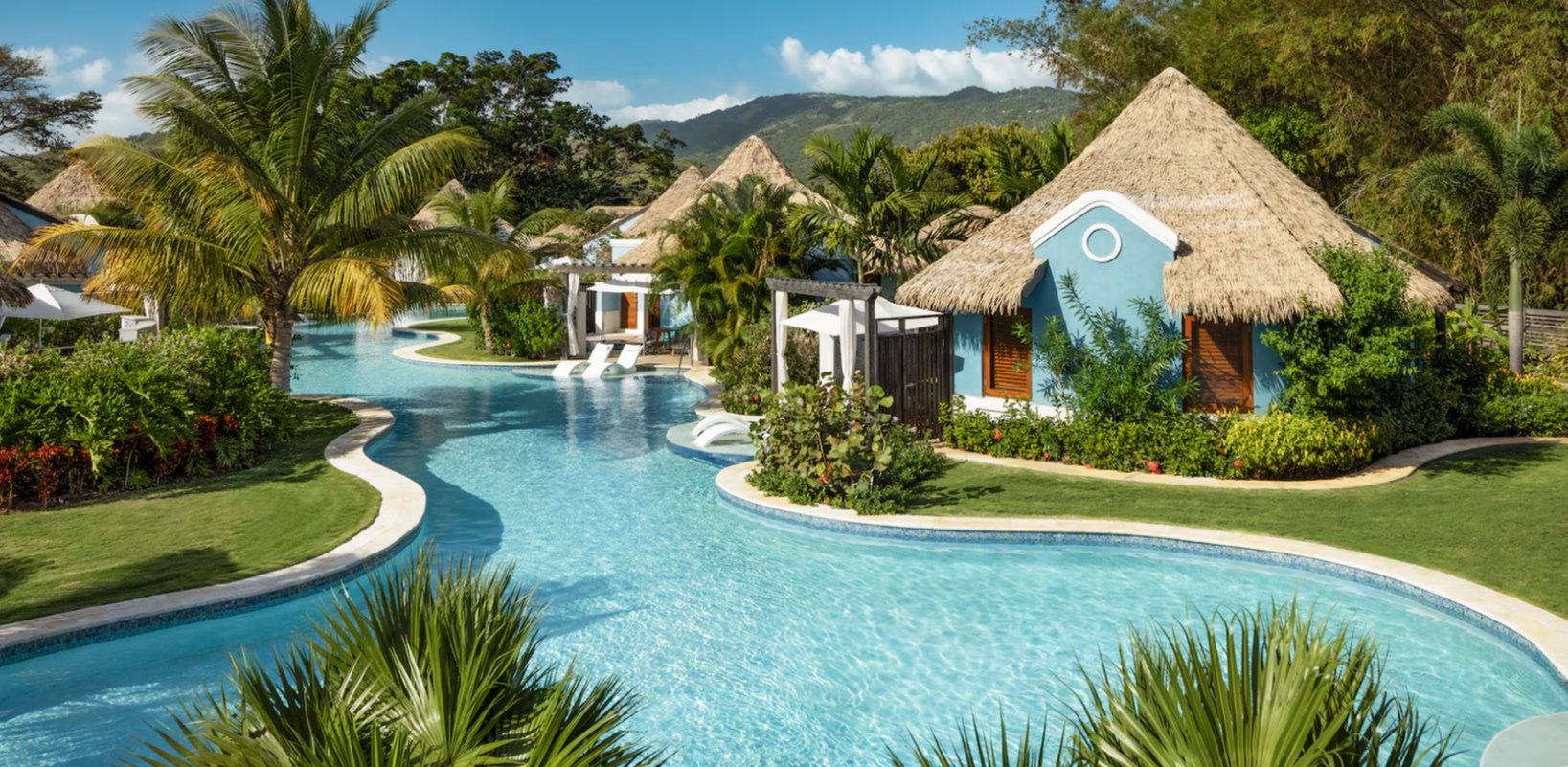Lush tropical resort with a pool, thatched-roof buildings, and green landscaping under a bright blue sky.