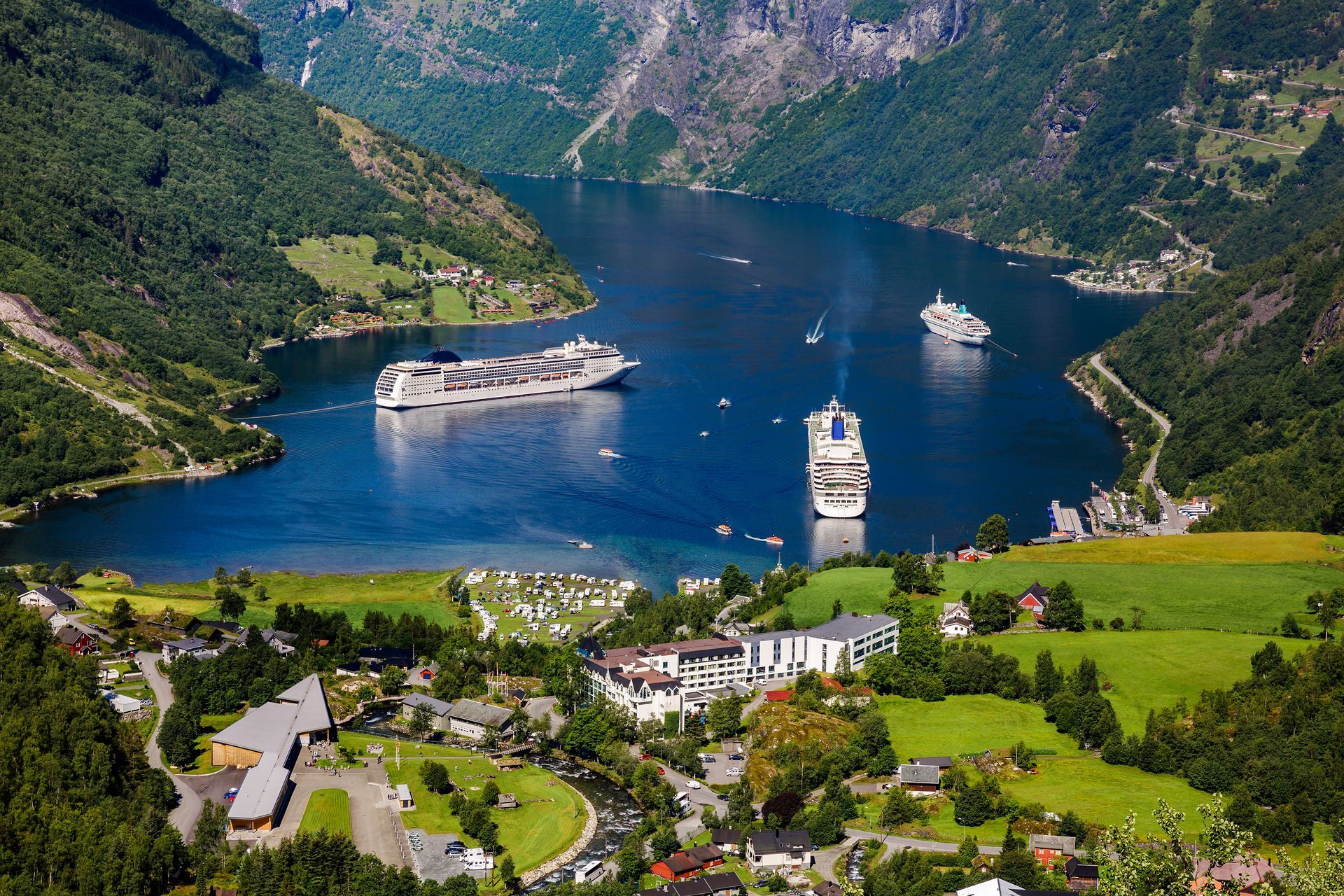 Aerial view of Geirangerfjord, Norway, with cruise ships on blue water, surrounded by green hills and a town.