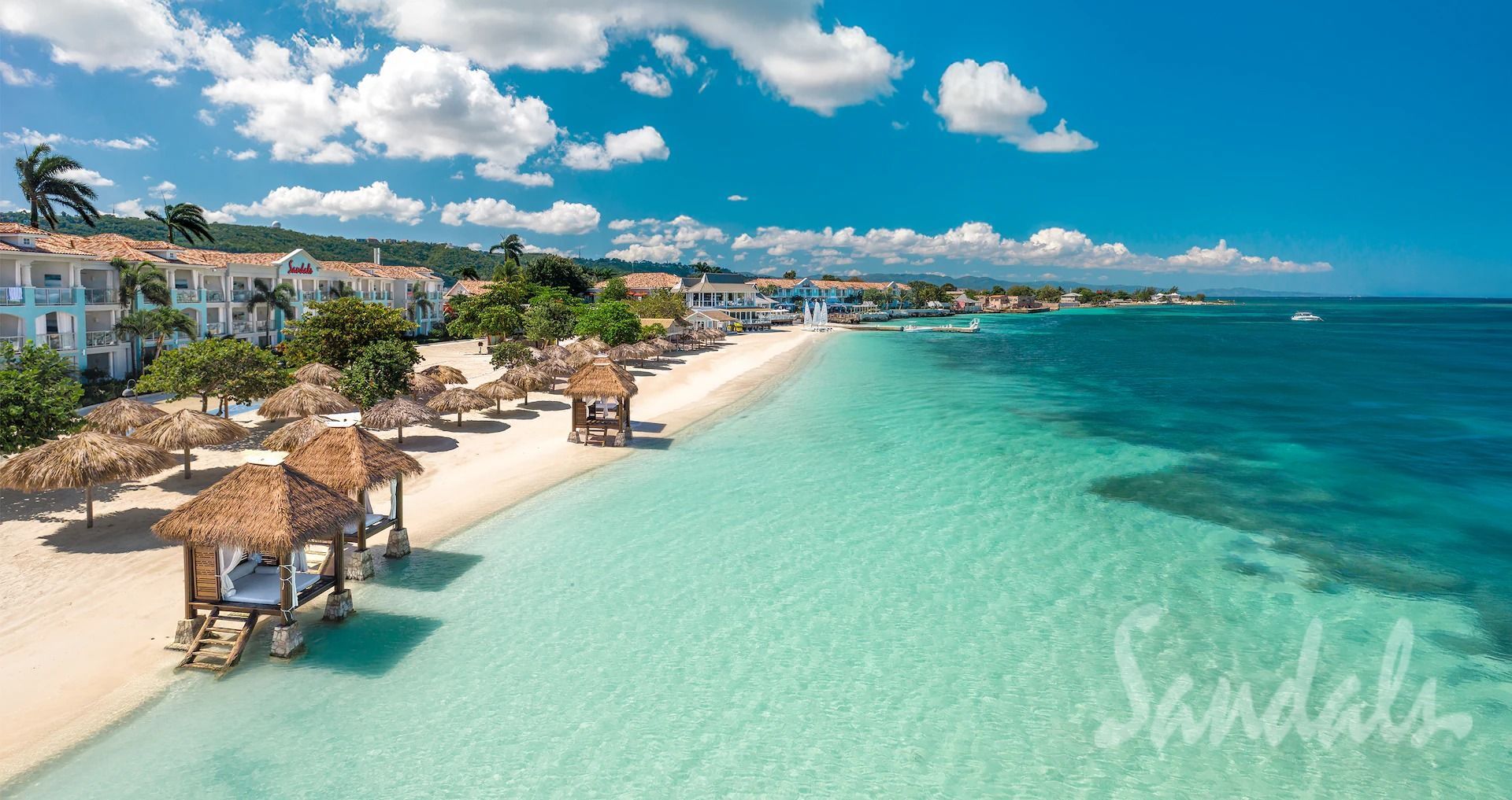Beach resort in Jamaica with turquoise water, white sand, thatched huts, and clear blue sky.