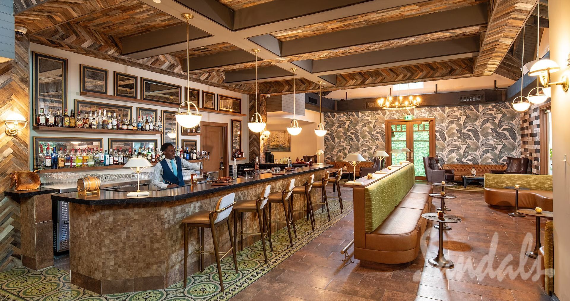 Inside a well-lit bar: bartender behind a textured counter; stools; green-carpeted floor; decorative ceiling; wall art.