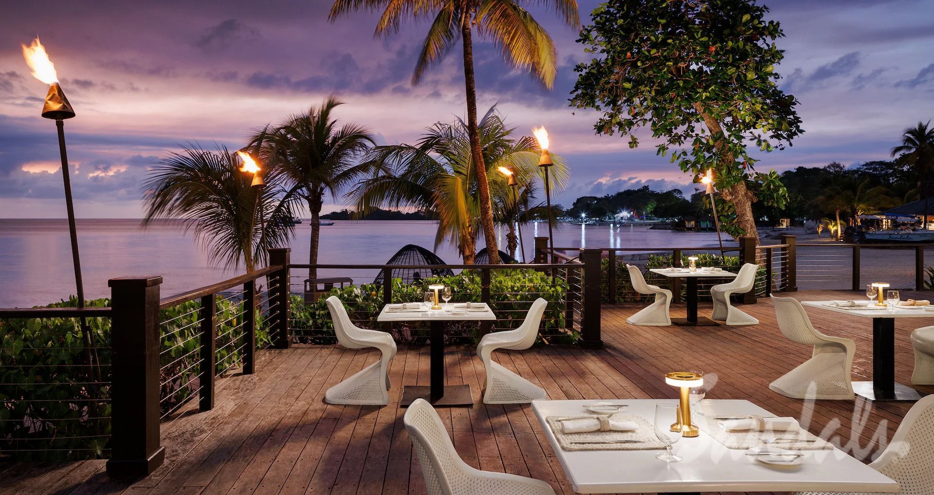 Restaurant patio overlooking the ocean at sunset. White chairs and tables, tiki torches, palm trees.