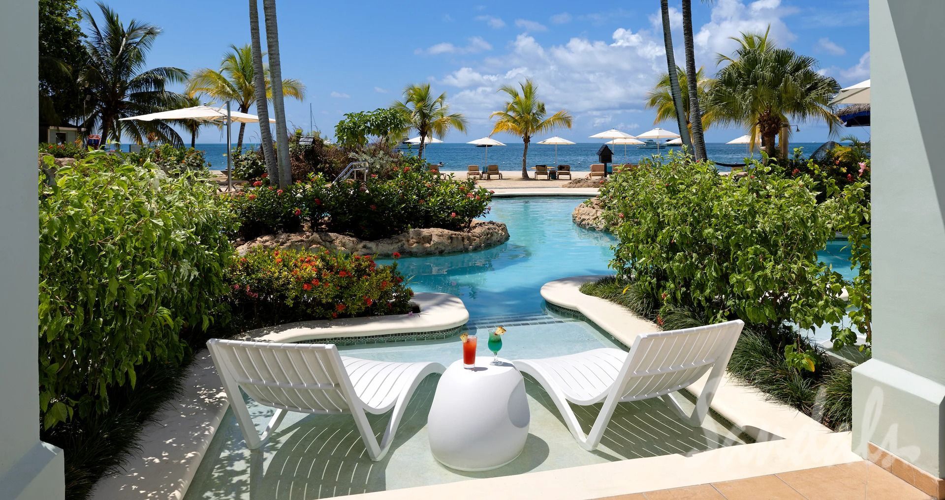 Poolside scene: white lounge chairs, cocktails, and ocean view in a tropical setting.