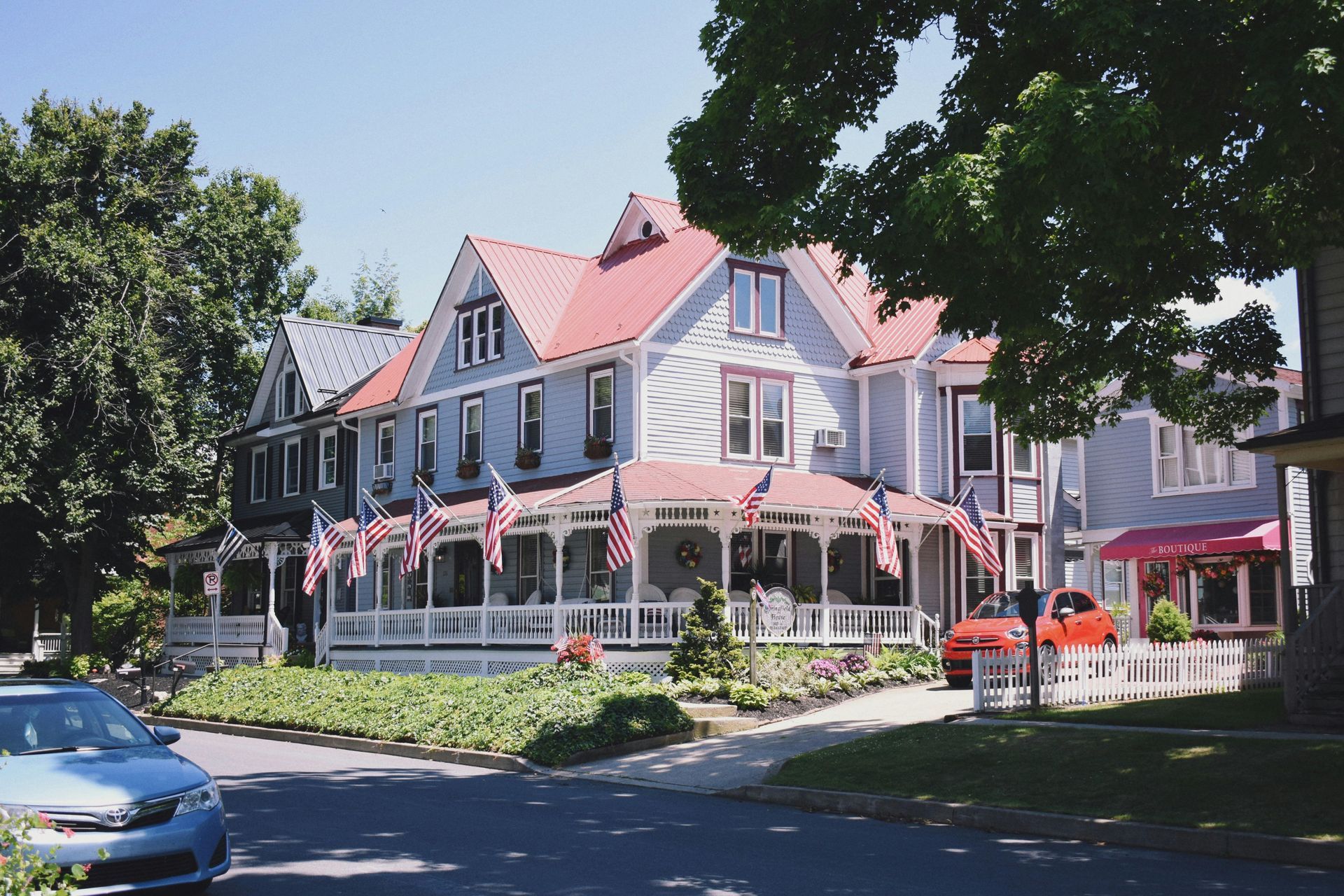 Victorian house with American flags, blue siding, red roof, and white porch. Sunny day, street view.