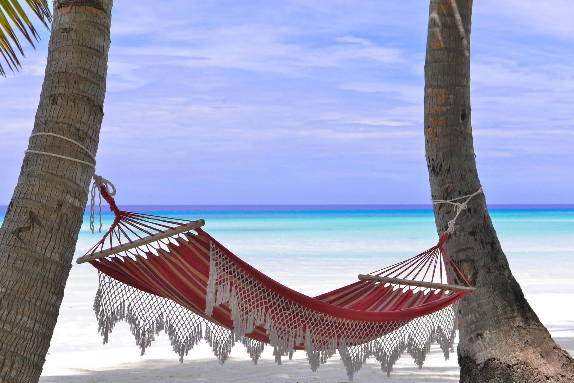 Red hammock strung between palm trees on a white sandy beach with a turquoise ocean in the background.