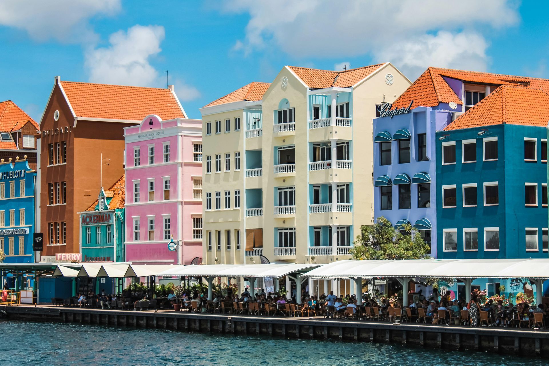 Colourful buildings  waterfront in Willemstad, Curaçao. People sit at tables under a white awning.