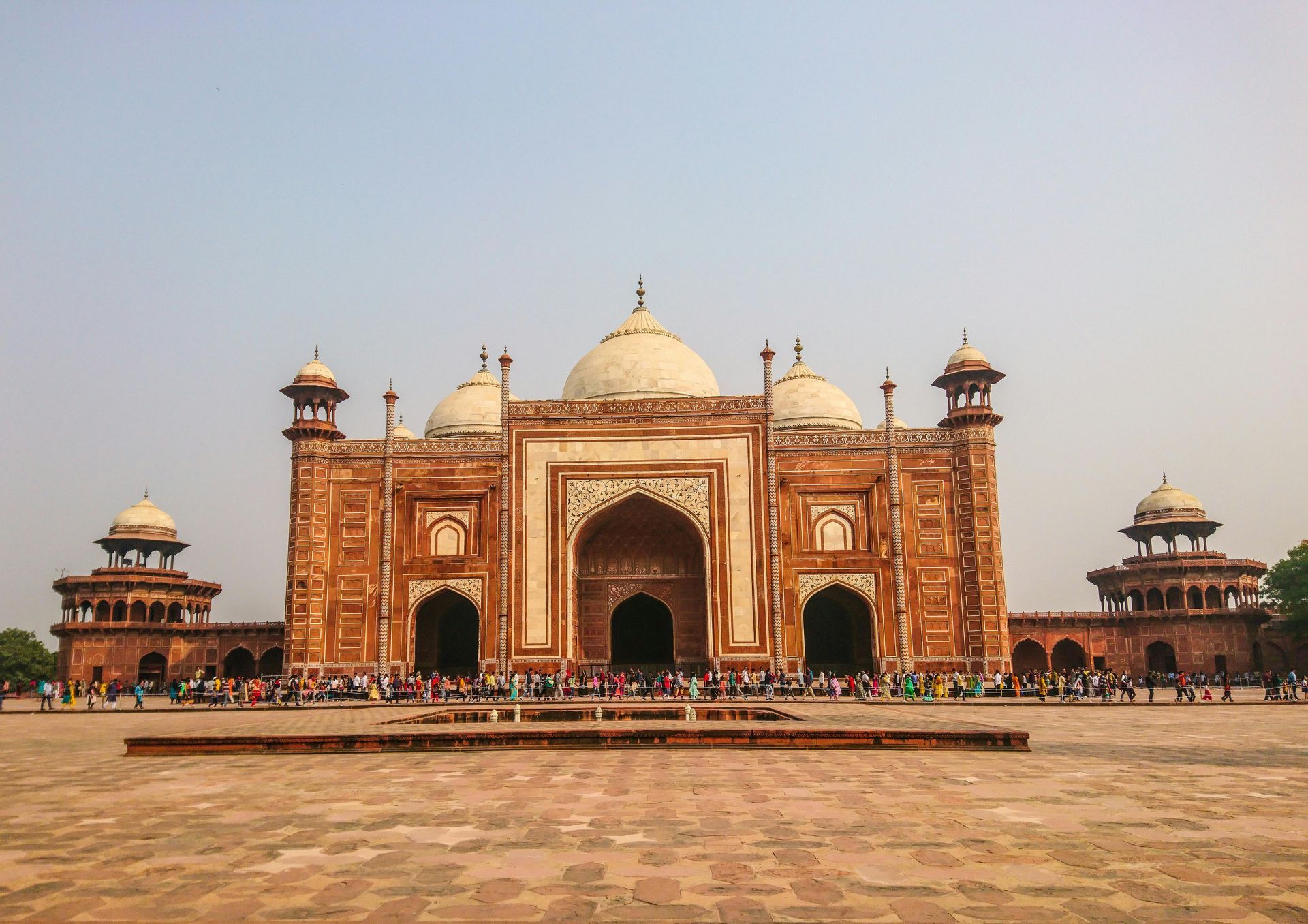 Monument with arches and domes; people gathered outside. Brick-colored building with white accents under a blue sky.
