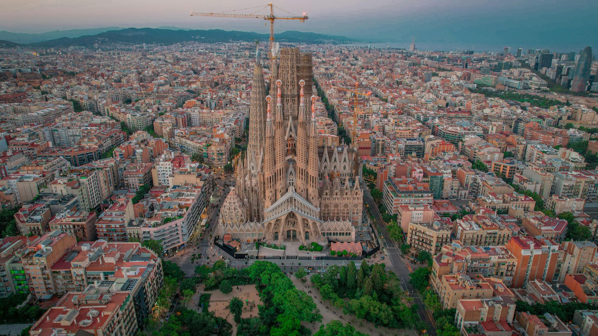 Aerial view of Barcelona's Sagrada Familia church, towering over a dense urban landscape, under a soft sky.
