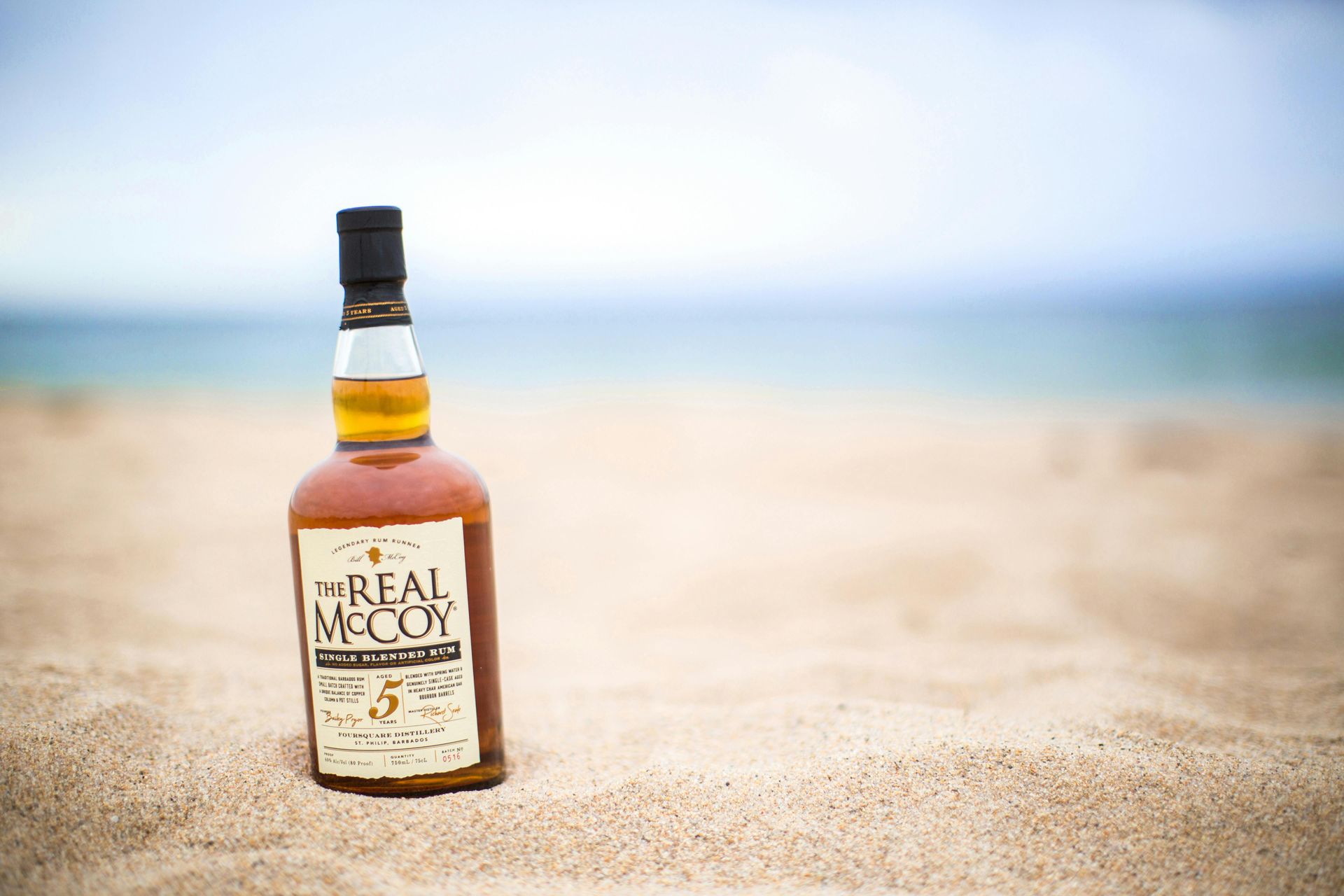 Bottle of The Real McCoy rum on a sandy beach with the ocean in the background.
