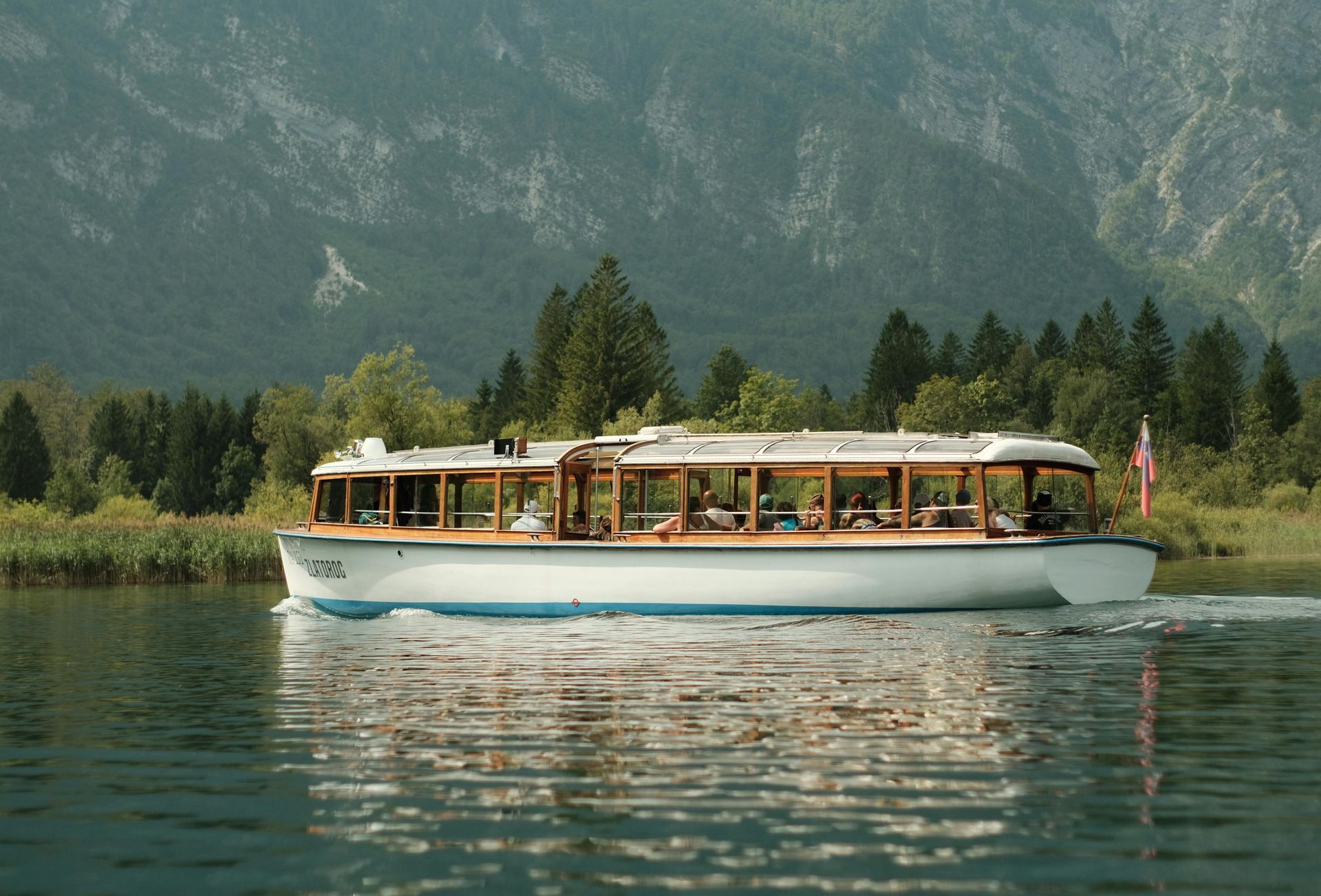 A boat with passengers on a lake, mountains and trees in the background.