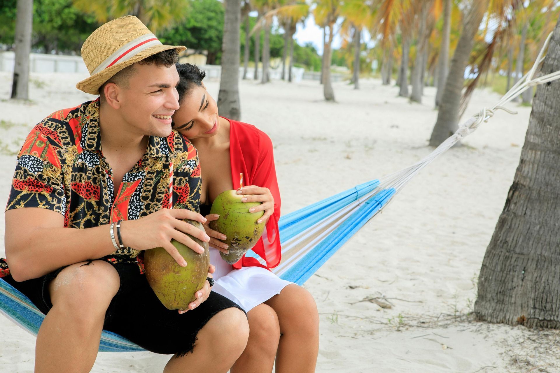 Couple relaxing in hammock on beach, drinking from coconuts; man wearing straw hat and patterned shirt.