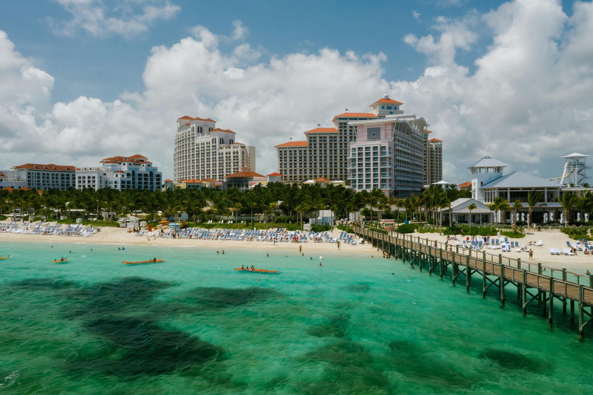 Beach scene with turquoise water, pier, resort buildings, and people on the sand.