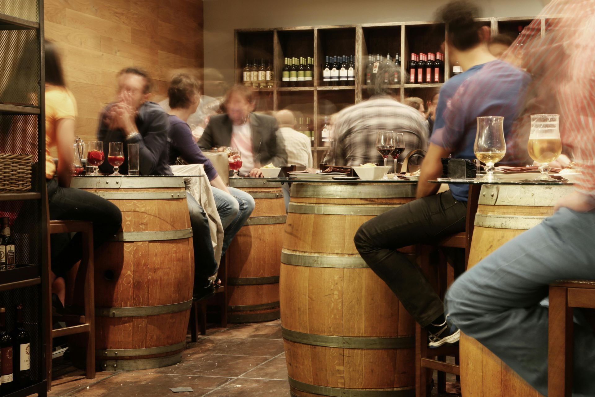 People in a wine bar, seated at barrel tables. Shelves of wine in the background, blurred motion.