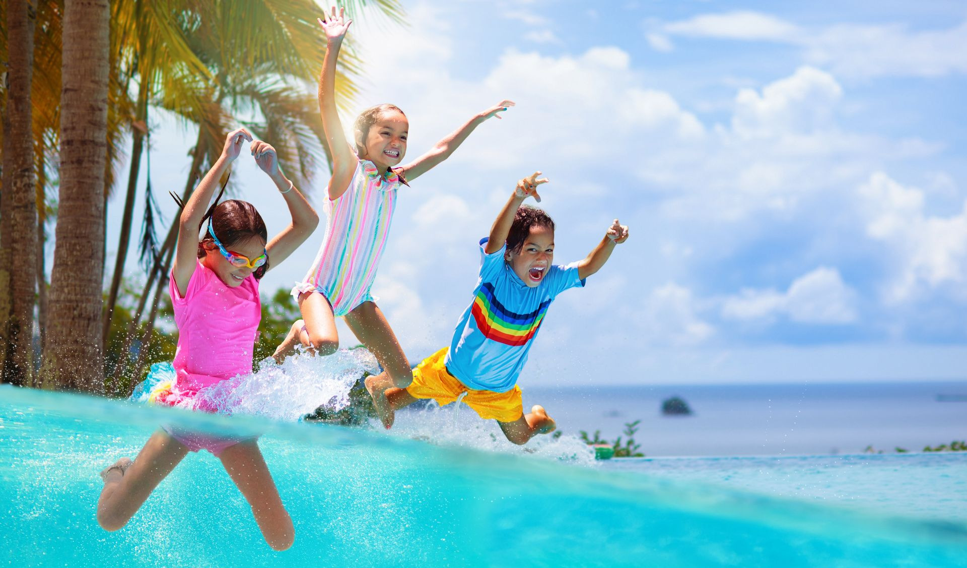 Three children jumping into a swimming pool with ocean in the background; sunny day.