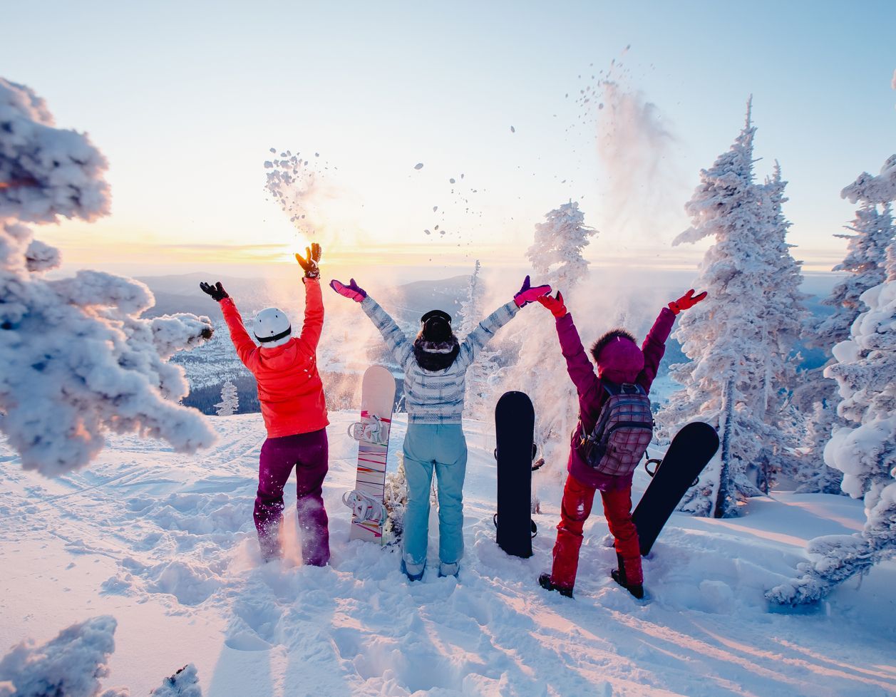 Three people with arms raised in a snowy landscape. Snowboards are beside them; trees are frosted.