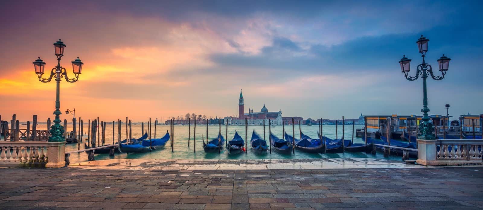 A row of gondolas are docked in a harbor at sunset.