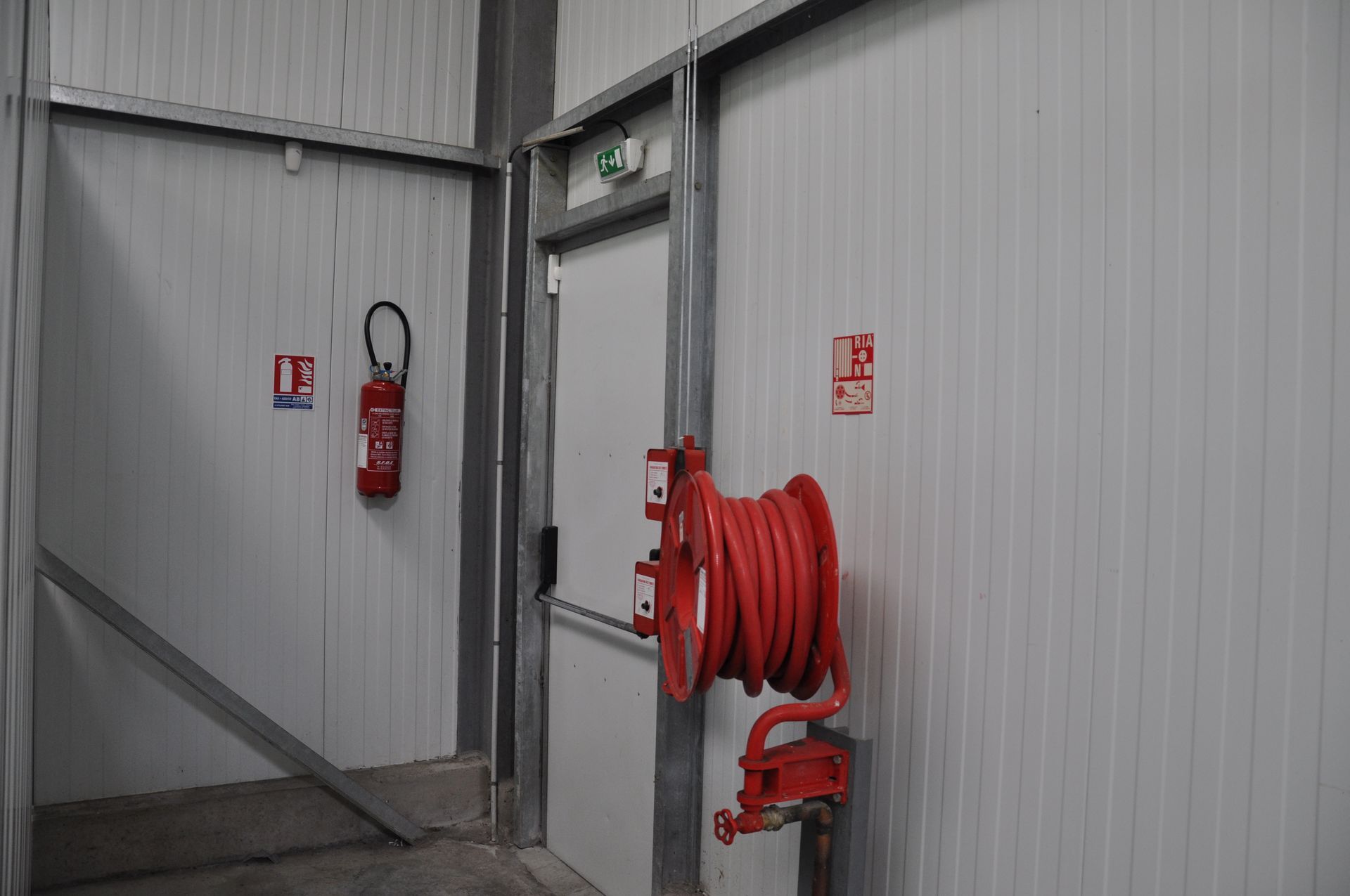 Équipements de sécurité incendie à l'intérieur d'un bâtiment aux murs blancs : un extincteur, un dévidoir de tuyau d'incendie et un panneau de sortie.