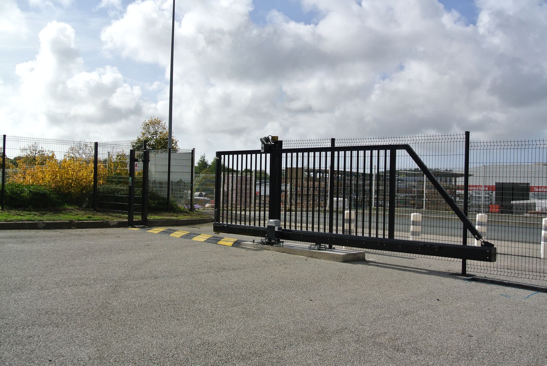 Portail de sécurité coulissant noir sur une surface d'asphalte grise ; clôture de barbelés en arrière-plan sous un ciel nuageux.