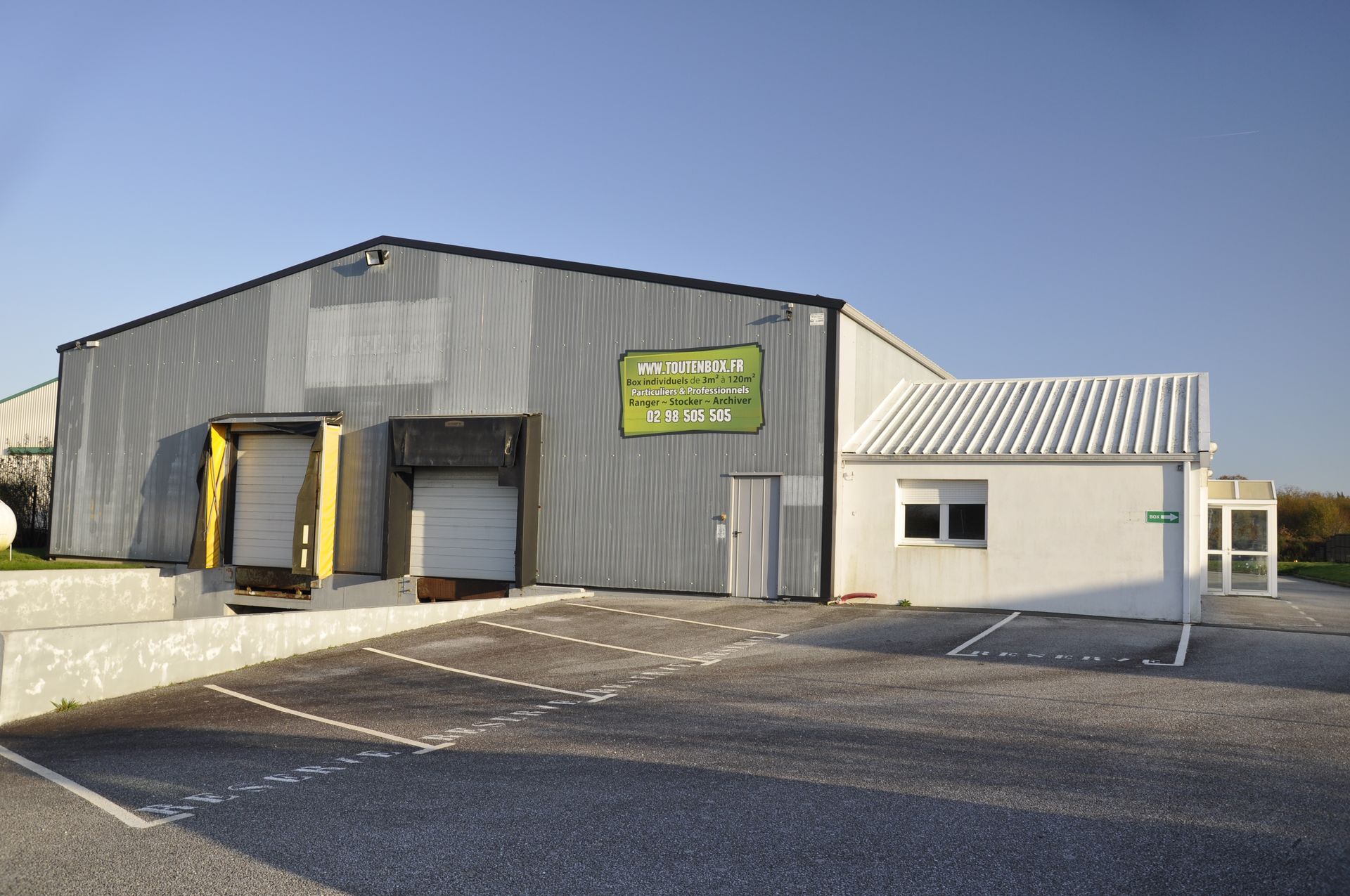 Entrepôt avec quais de chargement, petit bureau blanc, parking. Panneau « Produits frais de la ferme ».