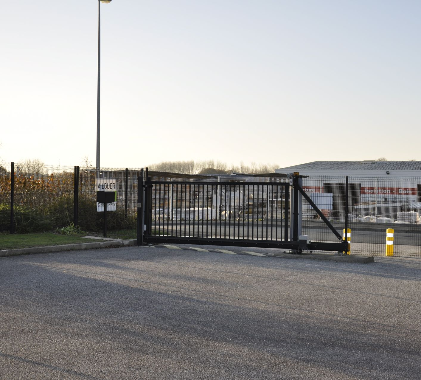 Portail noir, cadre industriel ; sol asphalté, clôture métallique, lampadaire et bâtiment en arrière-plan.