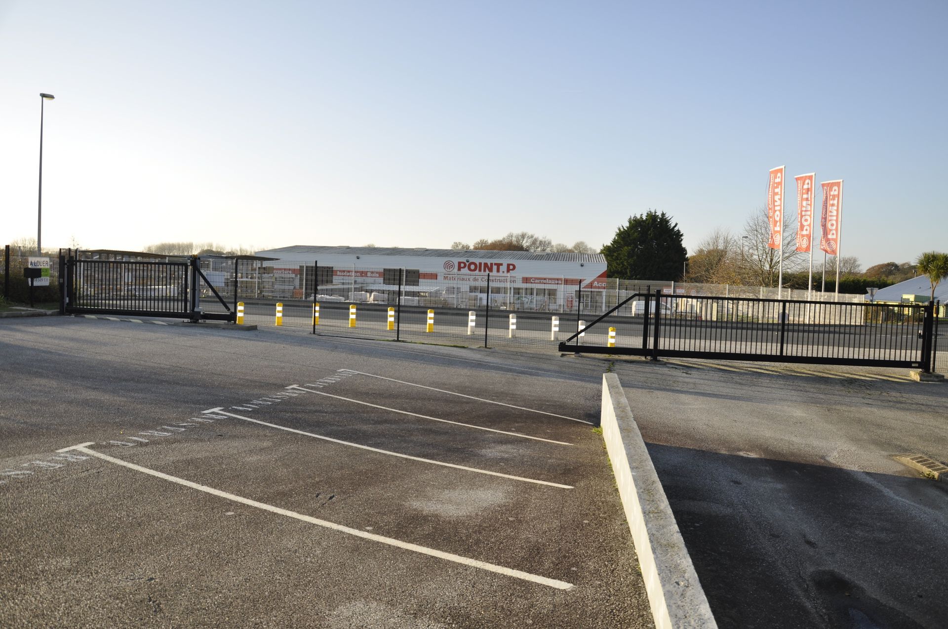 Des grilles noires s'ouvrent sur un parking et un bâtiment éloigné arborant une signalétique rouge.