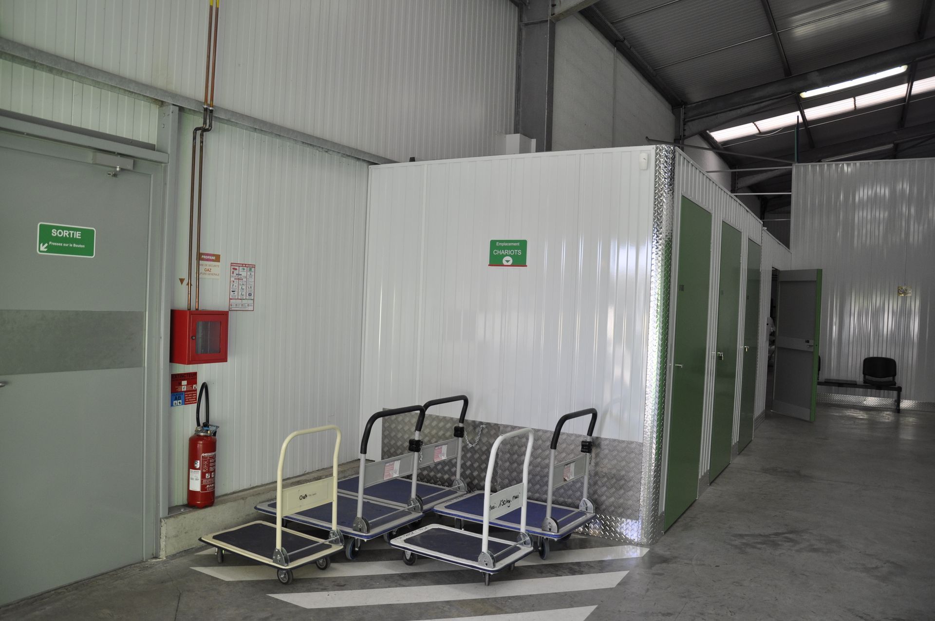 Unités de stockage avec portes sectionnelles vertes et parois en tôle ondulée. Sol en béton. Finitions blanches.
