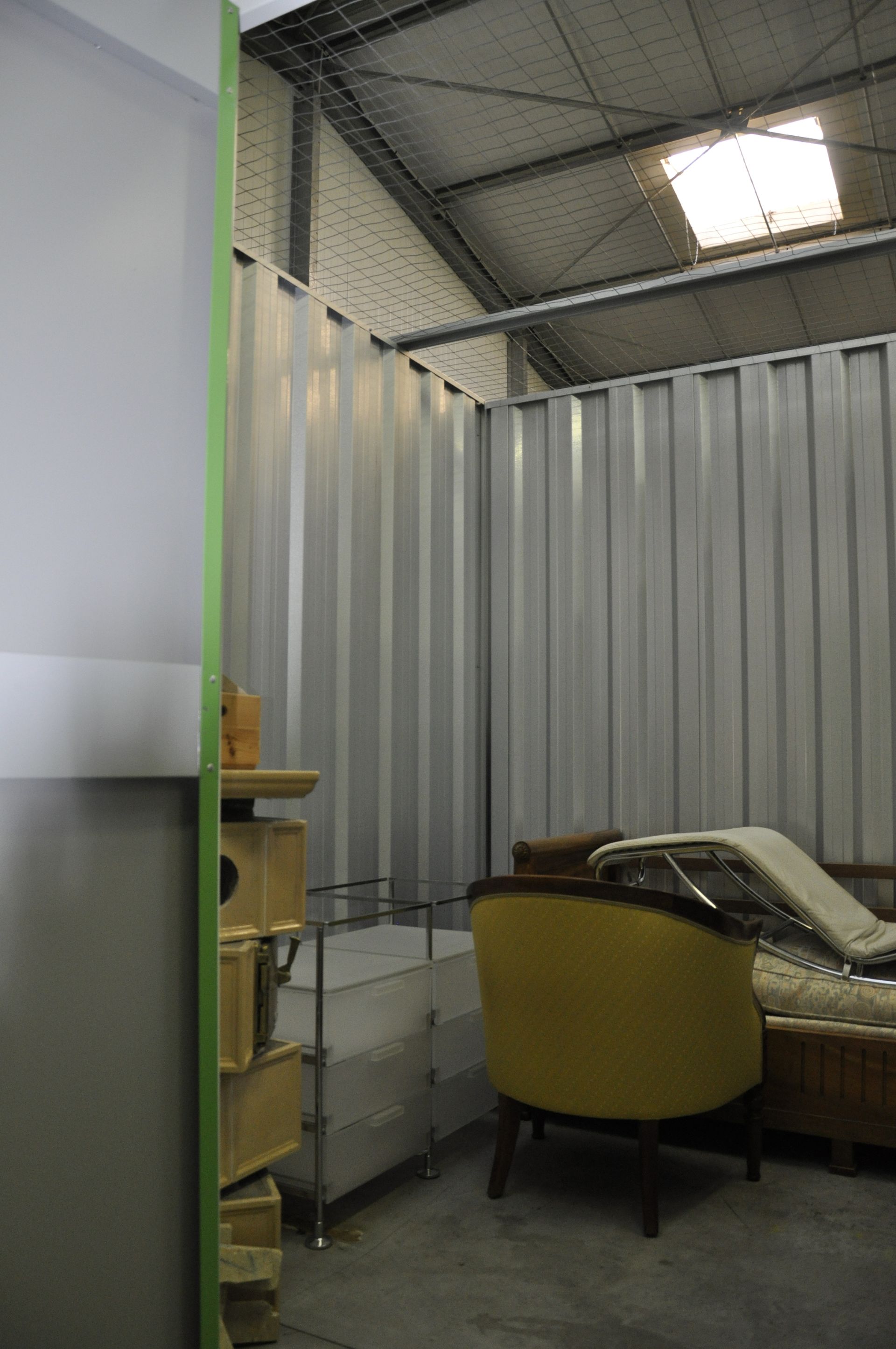 Intérieur aux parois en tôle ondulée, comprenant un lit et une chaise jaune. Teintes grises et vertes.