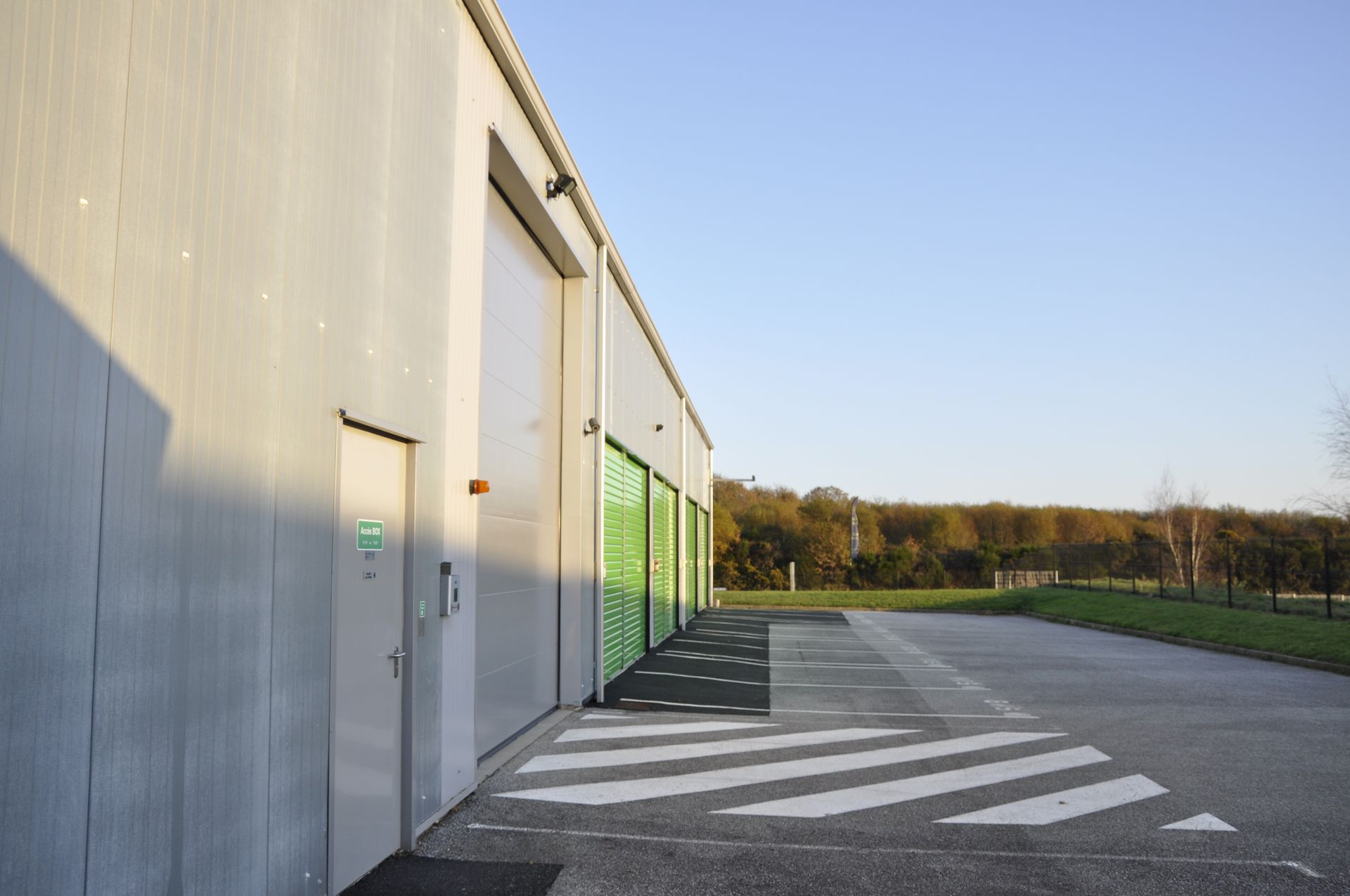 Extérieur d'un bâtiment commercial avec de grandes portes, un trottoir et une petite porte. Des portes vertes sont visibles. Ciel dégagé.