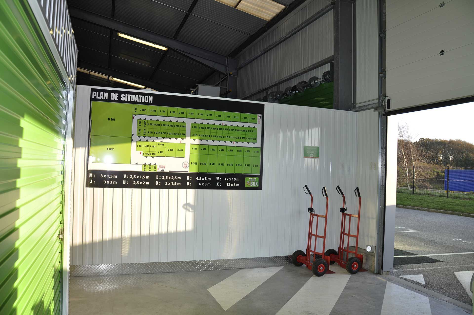 Unités de stockage avec portes sectionnelles vertes et parois en tôle ondulée. Sol en béton. Finitions blanches.