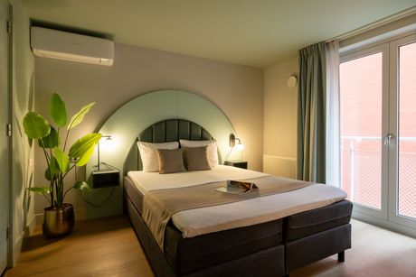 Een hotelkamer met een bed, decoratief hoofdeinde en een grote kamerplant. Groene muren, beige beddengoed en een balkondeur.