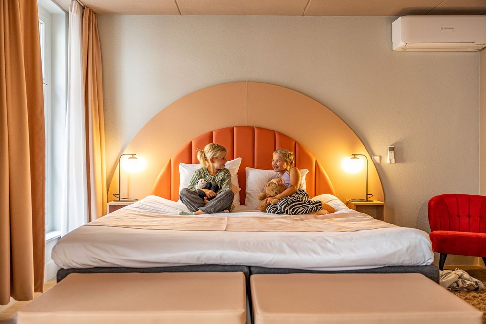 Twee jonge meisjes zitten op een groot bed in een hotelkamer. Het bed heeft een perzikkleurig hoofdeinde. Er staat een rode stoel naast.